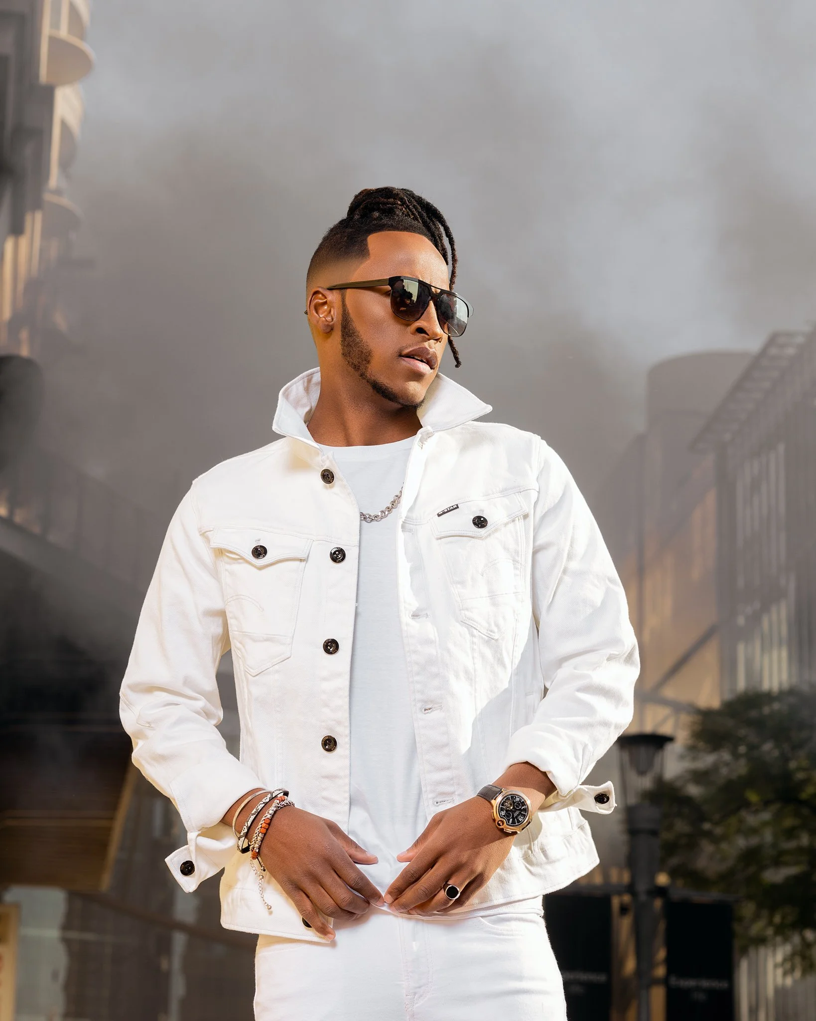 A stylish man wearing a white denim jacket, sunglasses, and accessories, standing outdoors with buildings and a smoky sky in the background.