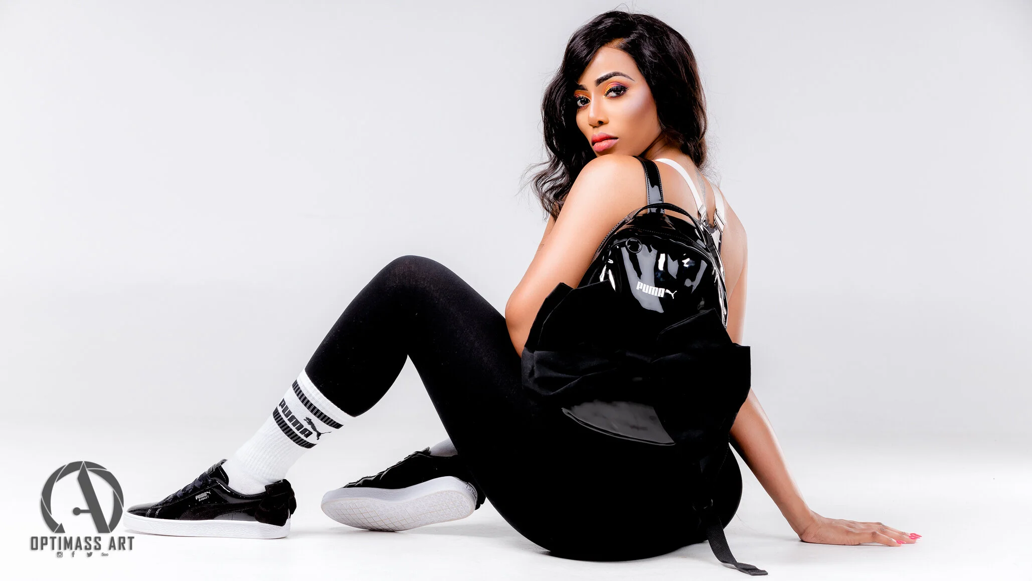 Young woman with long dark hair sitting on the floor with a black shiny Puma backpack, black leggings, white socks with Puma logo, and black sneakers, posing against a plain white background.