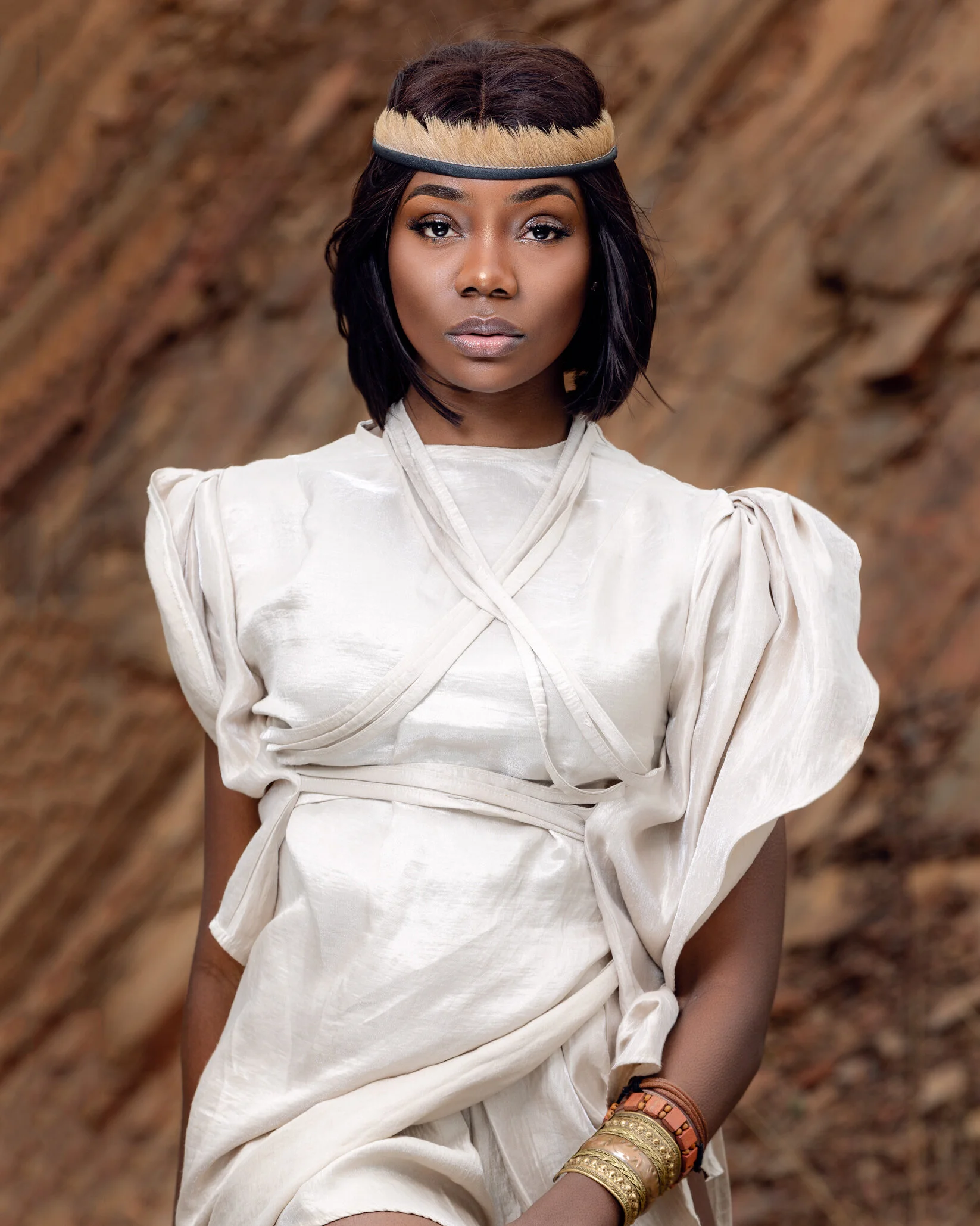A woman with dark hair wearing traditional African attire and jewelry, standing outdoors with a blurred natural background.