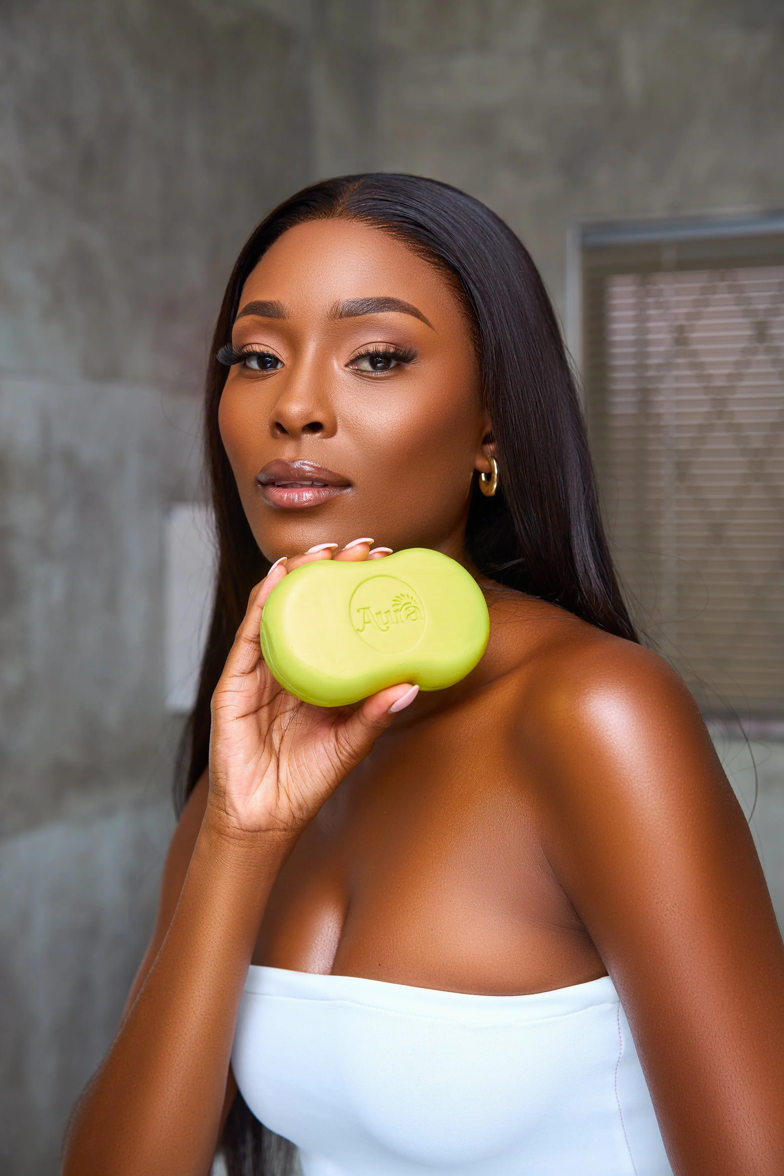 A woman with long black hair and gold hoop earrings holding a green bar of soap labeled 'Aura.' She is looking at the camera with a neutral expression.