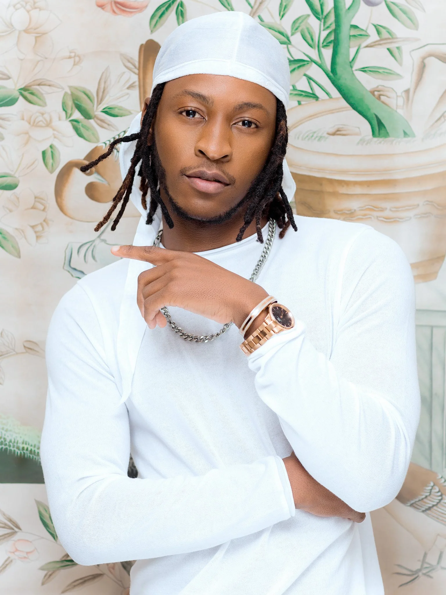 A young man with dreadlocks wearing a white bandana, white long-sleeve shirt, and gold watch, posing in front of a painted background with plants and a stone fountain.