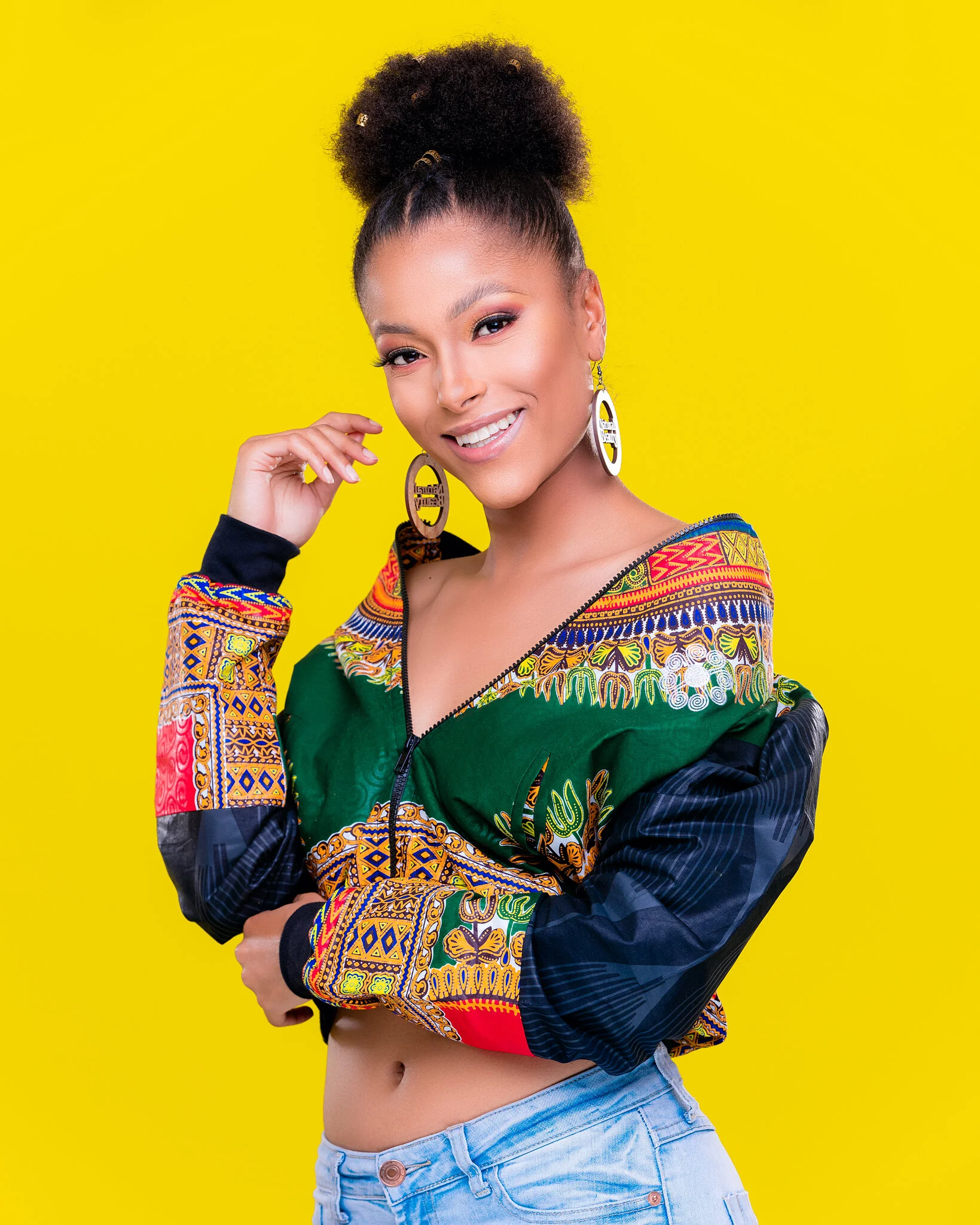 A woman with a curly high bun hairstyle, wearing large earrings, a colorful patterned jacket, and light blue jeans, standing against a bright yellow background.