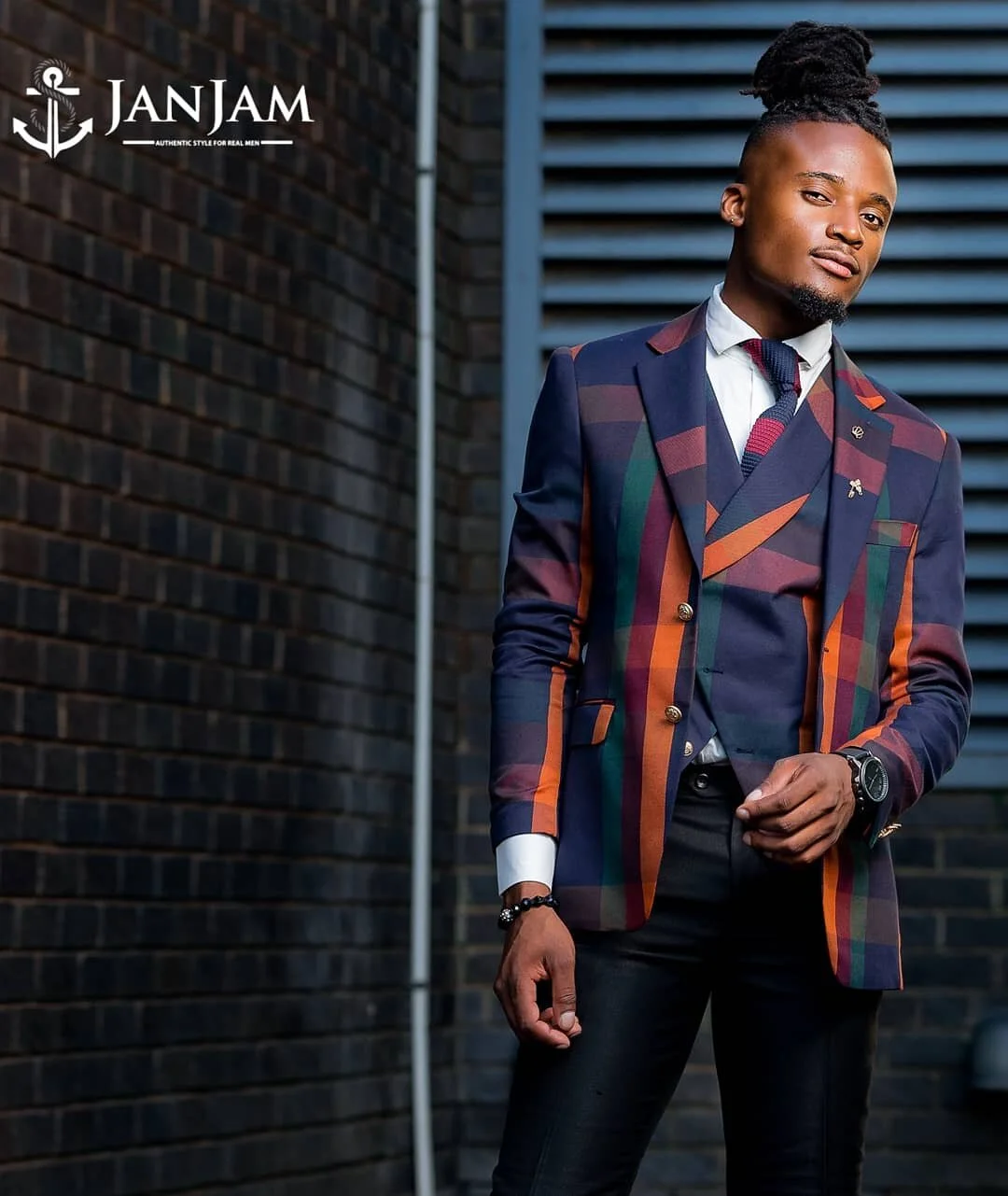 A stylish man with a high bun hairstyle wearing a colorful striped suit with a white shirt, a patterned tie, and black pants standing outdoors against a brick wall and blue louvered panels.