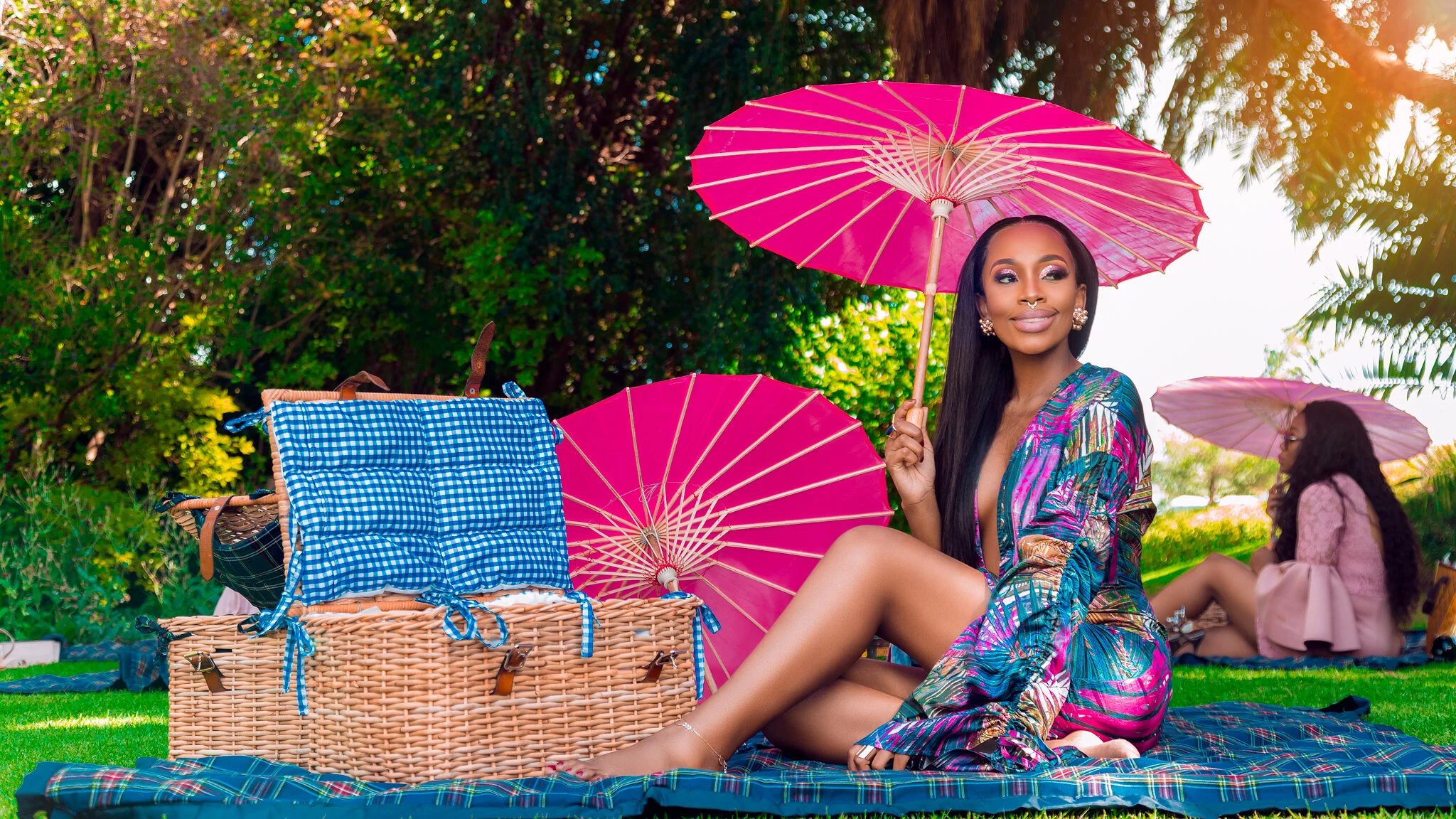 A woman sitting on a picnic blanket outdoors holding a pink umbrella, with a picnic basket and another pink umbrella nearby, while a girl relaxes in the background under another pink umbrella, surrounded by greenery.