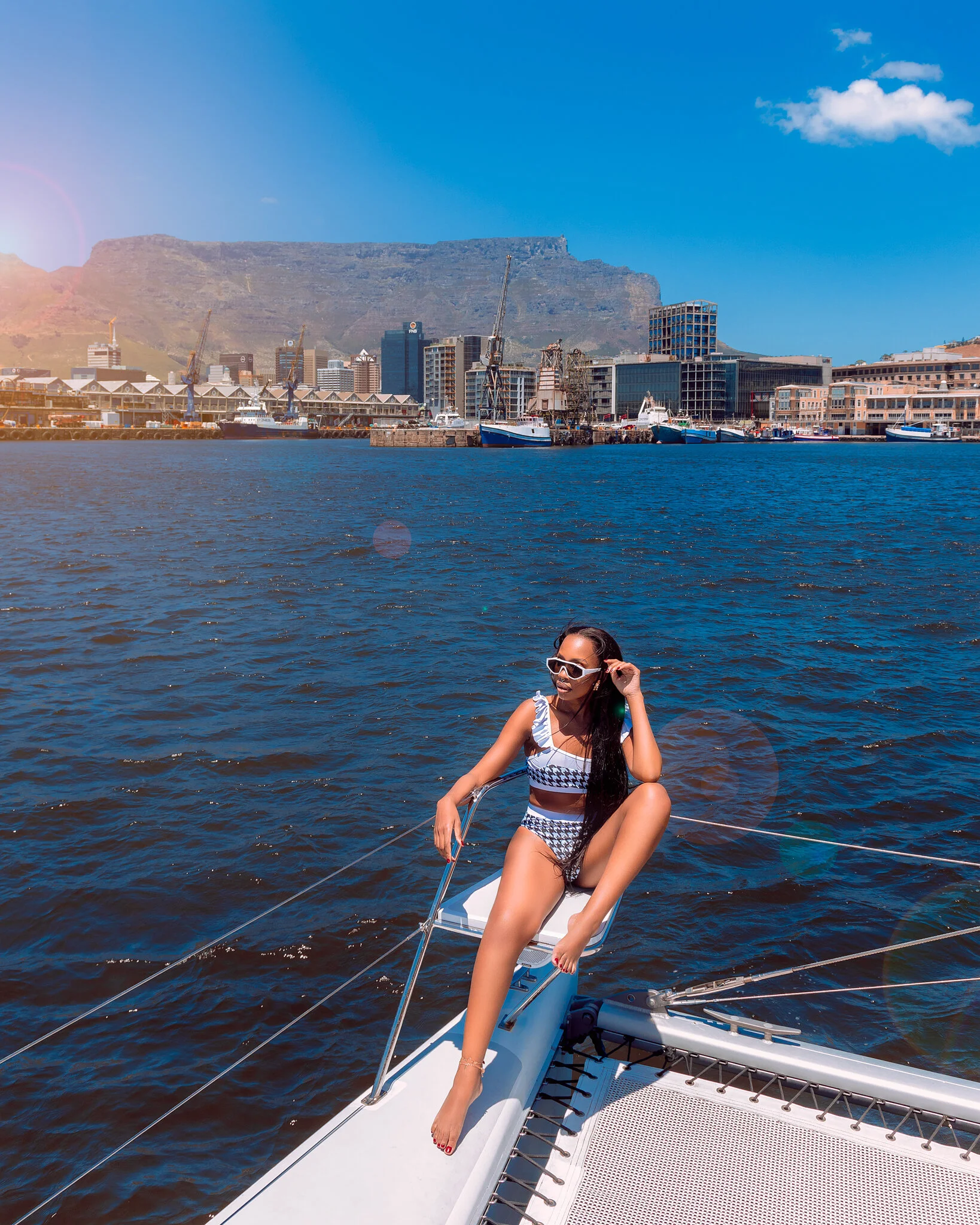 A woman in a striped bikini and sunglasses relaxing on the edge of a sailboat on a sunny day, with a city skyline, boats, and a mountain in the background.