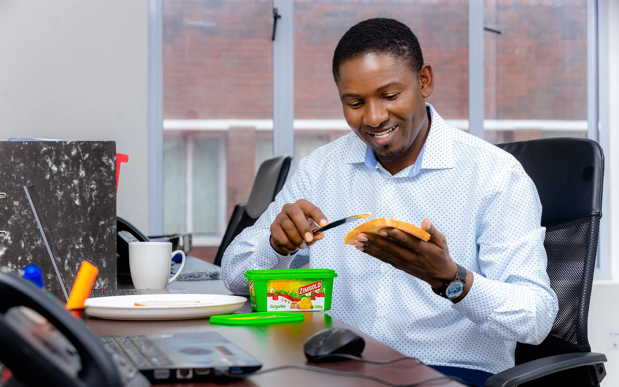A man sitting at a desk in an office, spreading margarine on bread with a butter knife. There is a green tub of margarine in front of him and office supplies on the desk.
