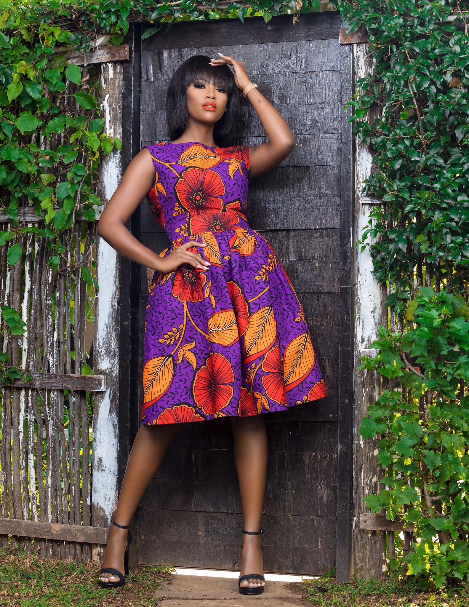 A woman in a colorful, sleeveless, floral dress standing in front of a wooden fence and greenery, striking a confident pose with one hand on her forehead and the other on her waist, wearing black high heels.