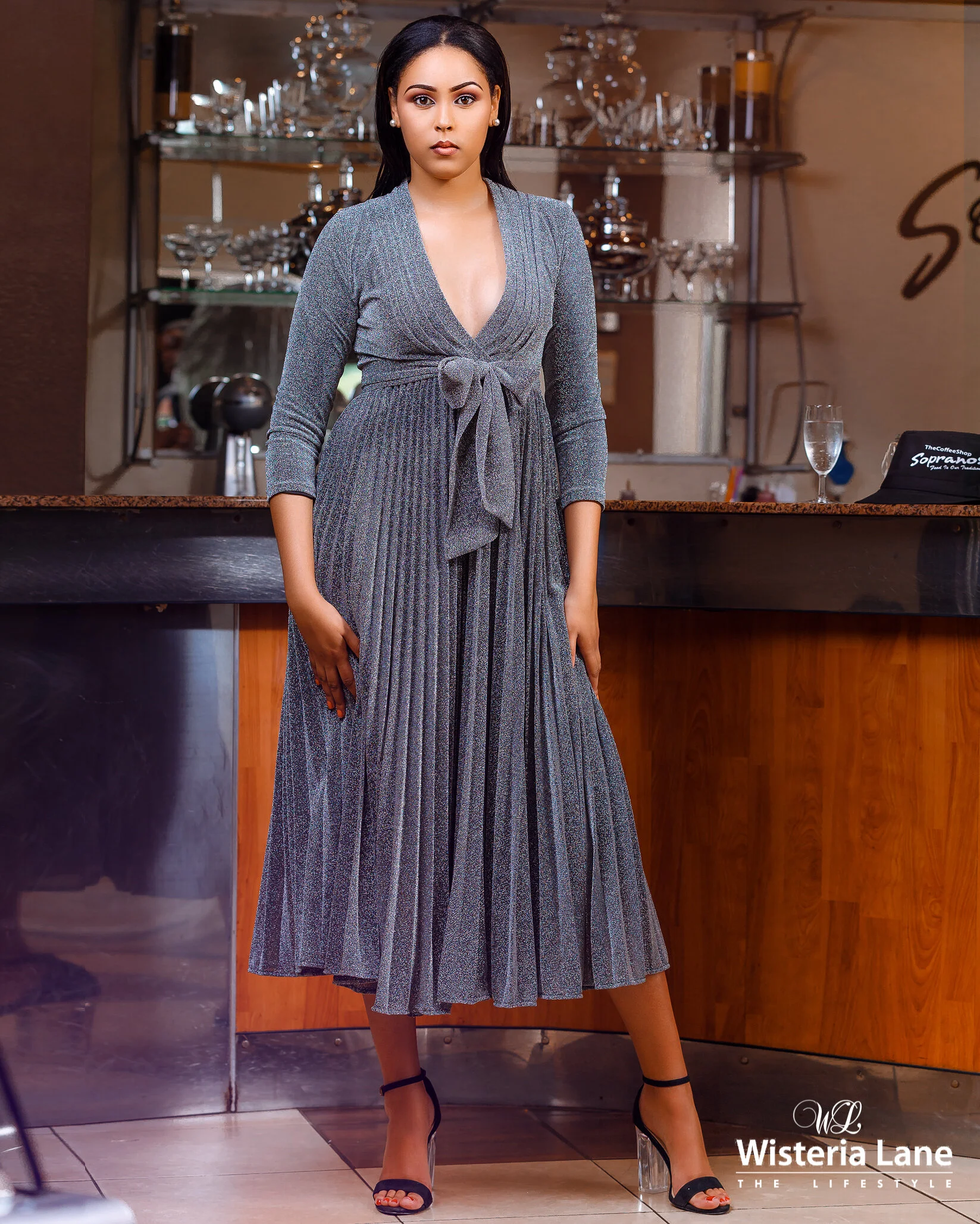 A woman stands behind a bar in a restaurant or cafe. She is wearing a long, gray, pleated dress with a tie at the waist and black high heels. The background shows glassware on shelves, a glass of water, and a nameplate on the bar.