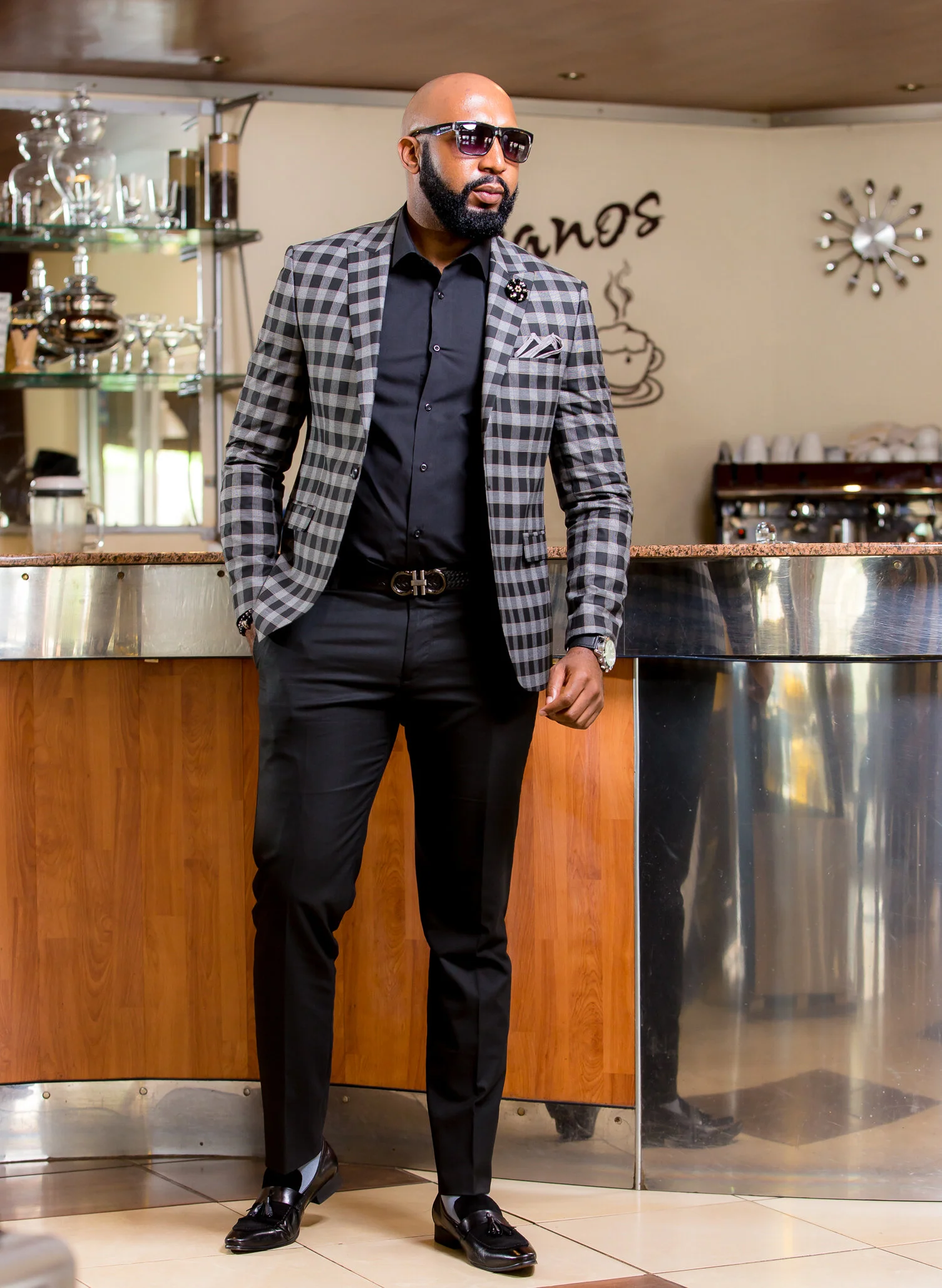 Stylish man in sunglasses, plaid blazer, and black dress shoes standing in a cafe or restaurant with a wooden counter.