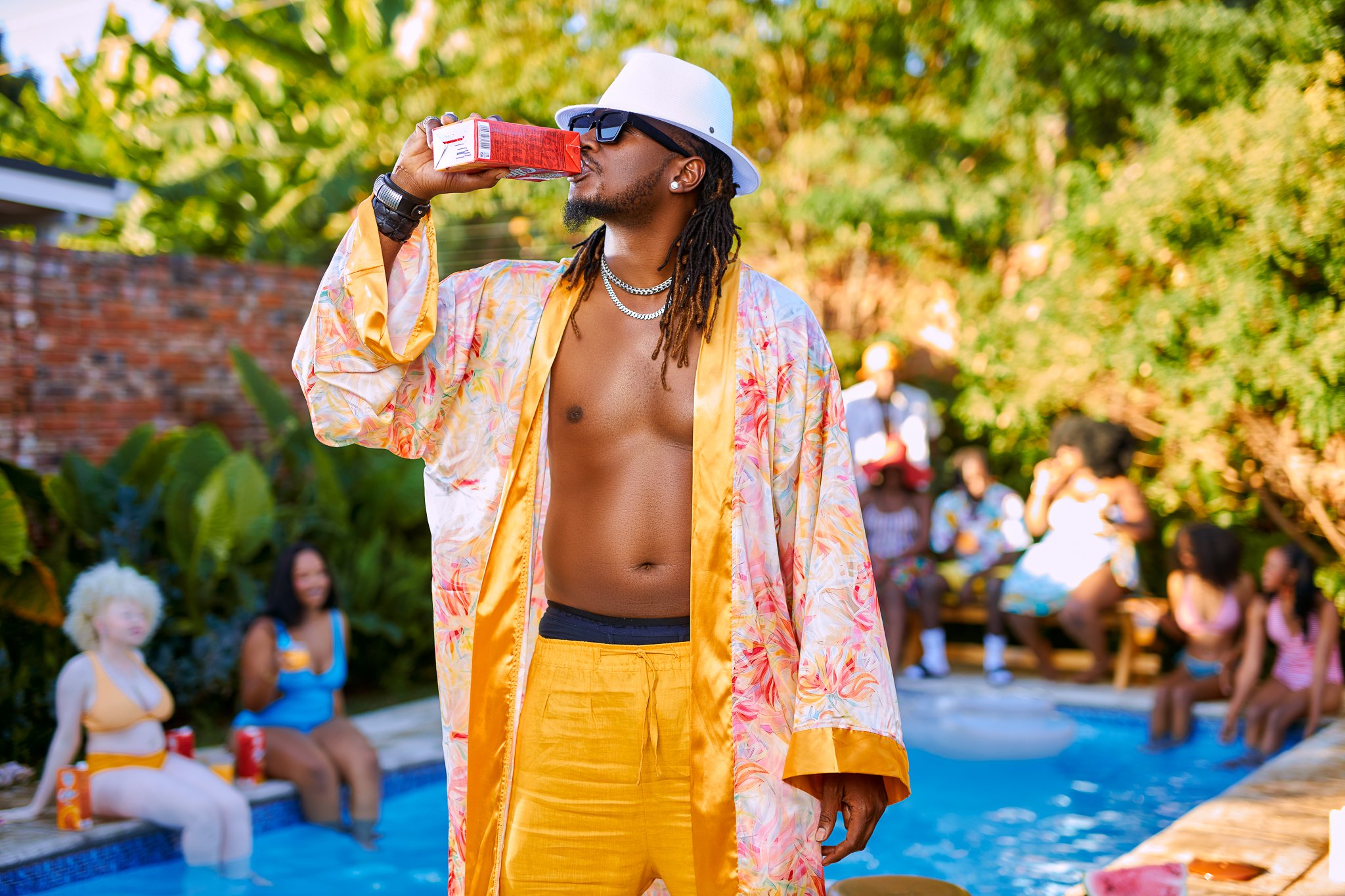 A shirtless man wearing a white hat, sunglasses, yellow shorts, and a floral robe is drinking from a juice box at a pool party with people sitting on the edge and in the background.