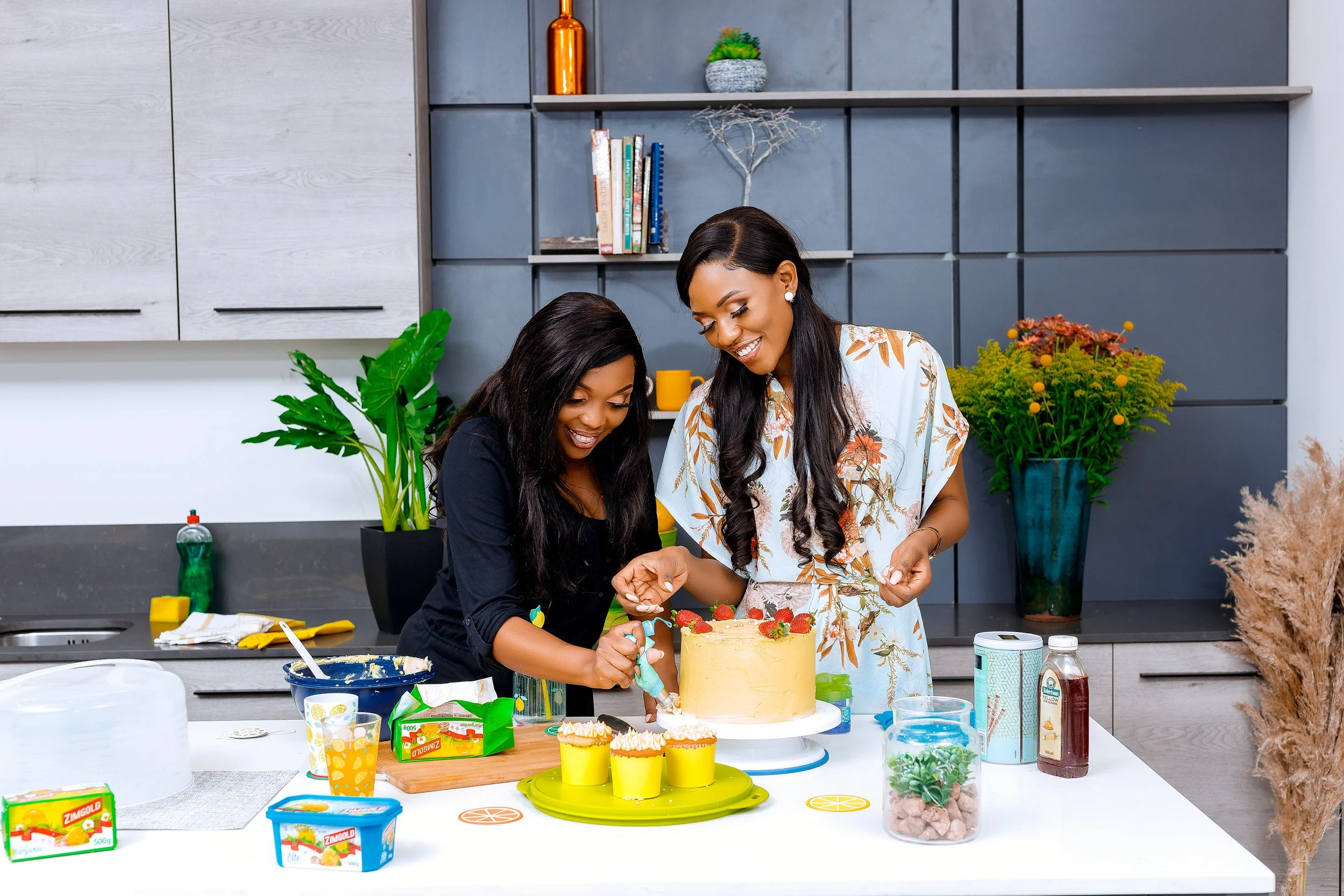 Two women are decorating a birthday cake in a modern kitchen. One woman is holding a piping bag, and they are both smiling. The counter has various food items, including cupcakes, frosting, and other ingredients.