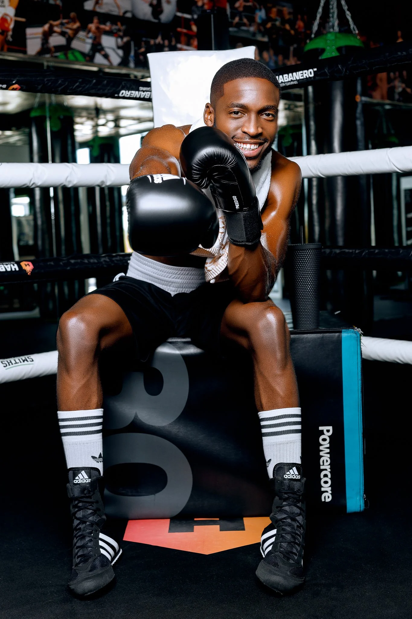 A young male boxer wearing black boxing gloves, black shorts, and white socks with black stripes, sitting in a boxing ring and smiling at the camera.
