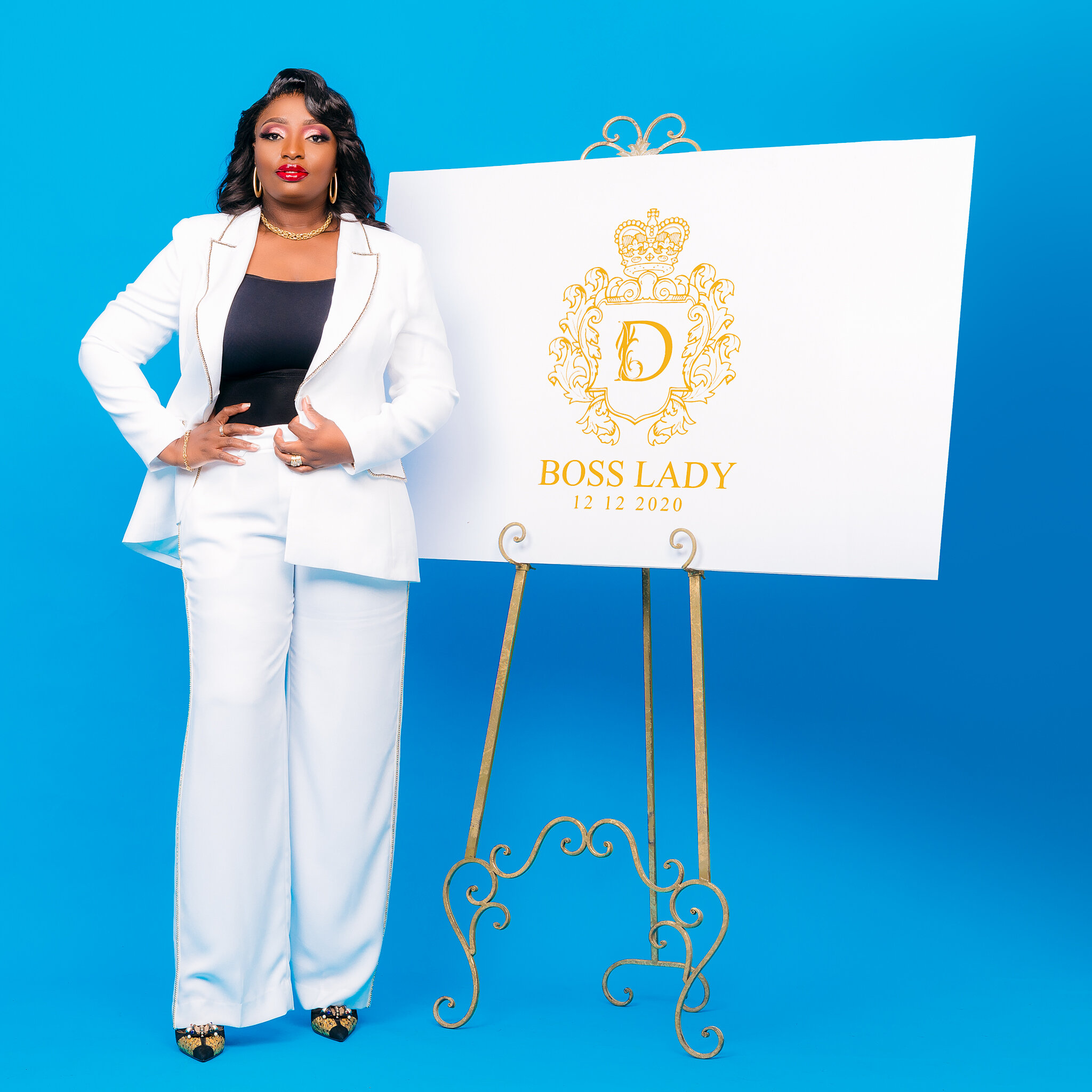 A woman dressed in a white suit with a black top, standing next to a sign that reads "Boss Lady" with a date of 12/12/2020. The sign has a decorative gold crest with a crown and a large letter D in the center.