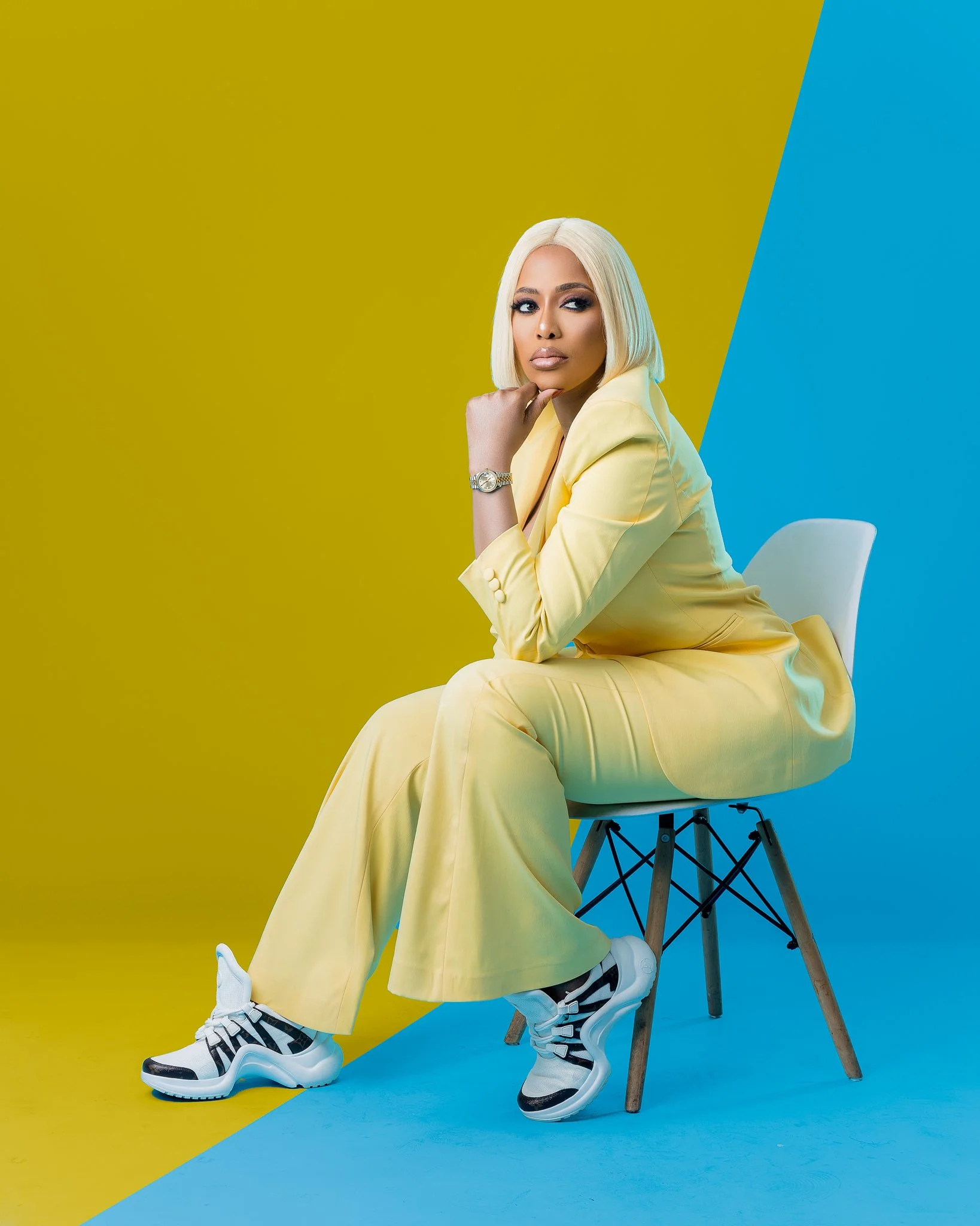 A woman with platinum blonde hair sitting on a modern white chair. She is dressed in a yellow suit, white sneakers, and wears a watch. The background features a colorful split of yellow and blue.