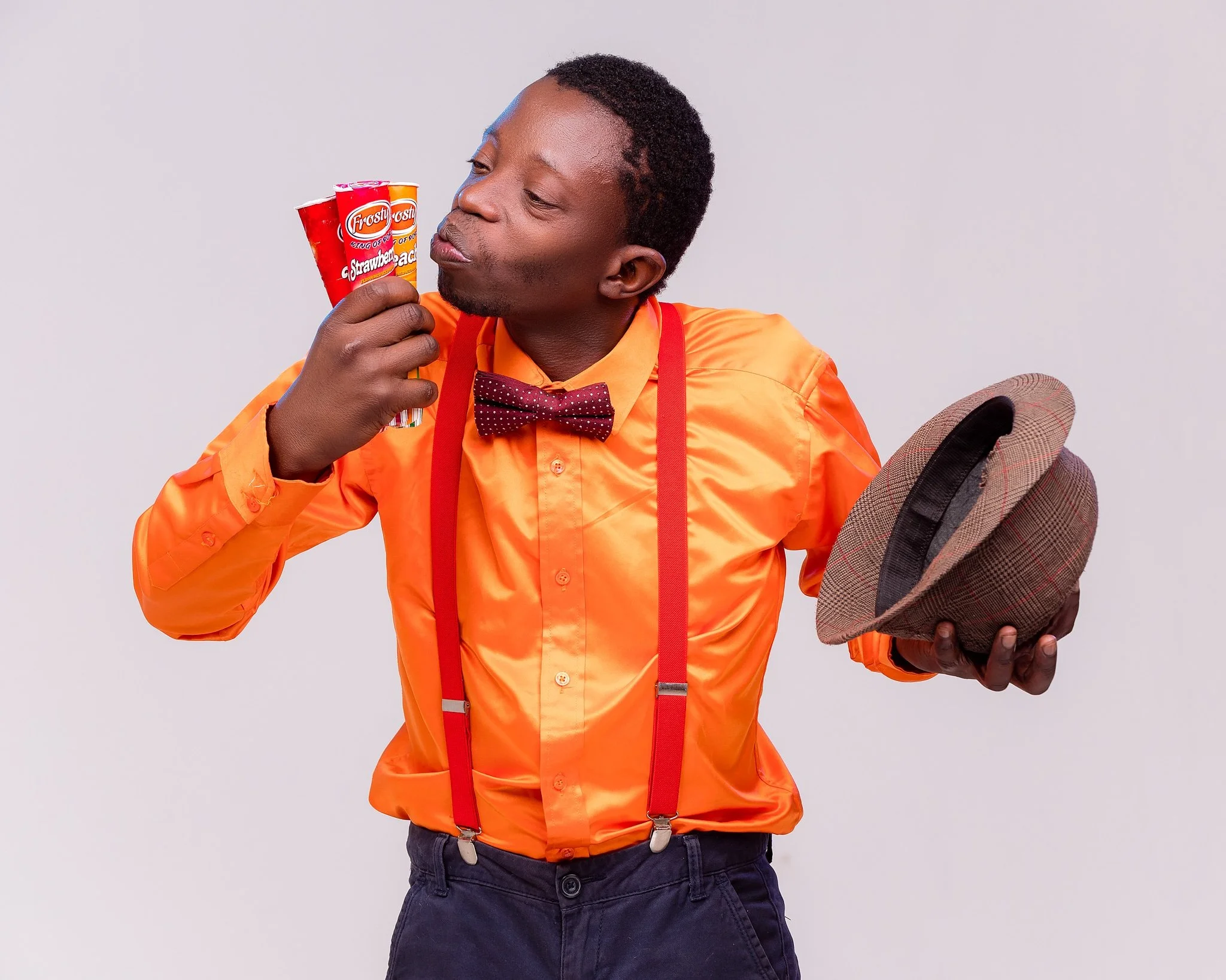 Man in orange shirt with suspenders and bowtie holding a hat in one hand and eating strawberry-flavored Frosty cups with the other.