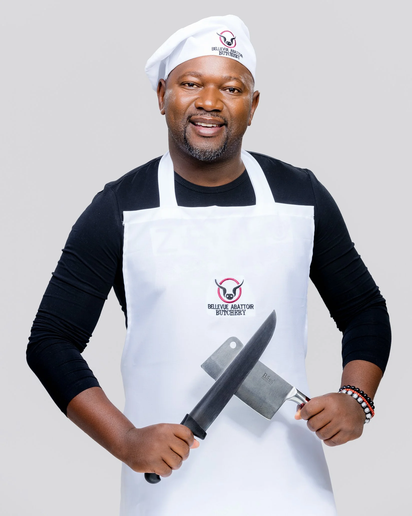 A man wearing a white chef's hat and apron, holding a meat cleaver and a knife, smiling in front of a plain background.