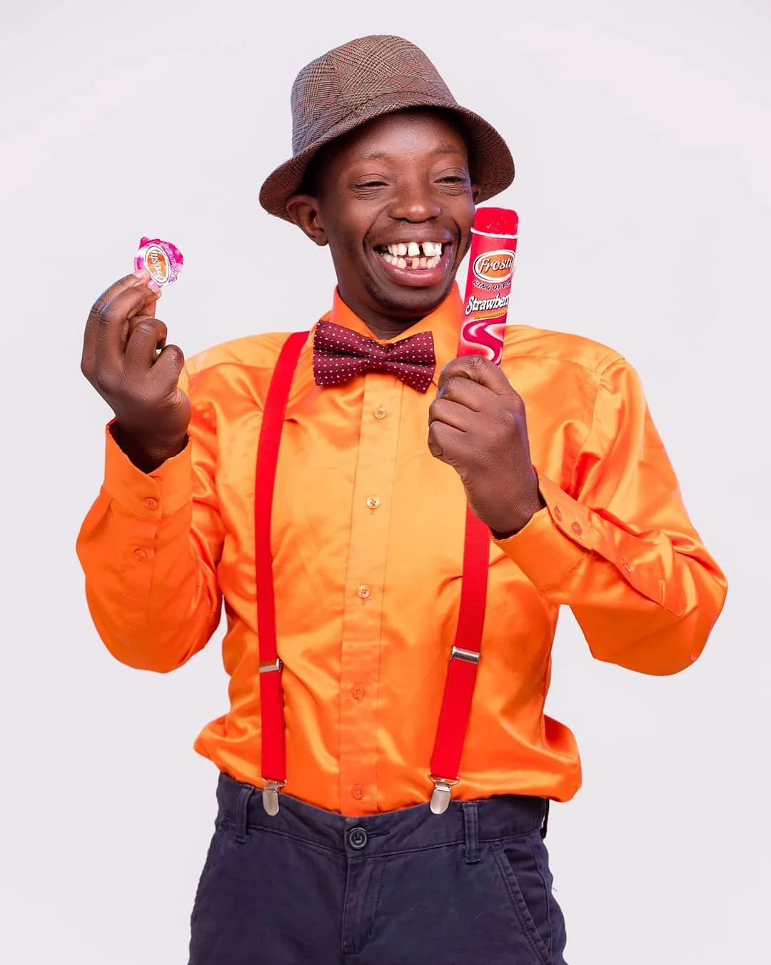 Smiling man in orange shirt, suspenders, and bowtie holding a strawberry-flavored ice cream and a lollipop.