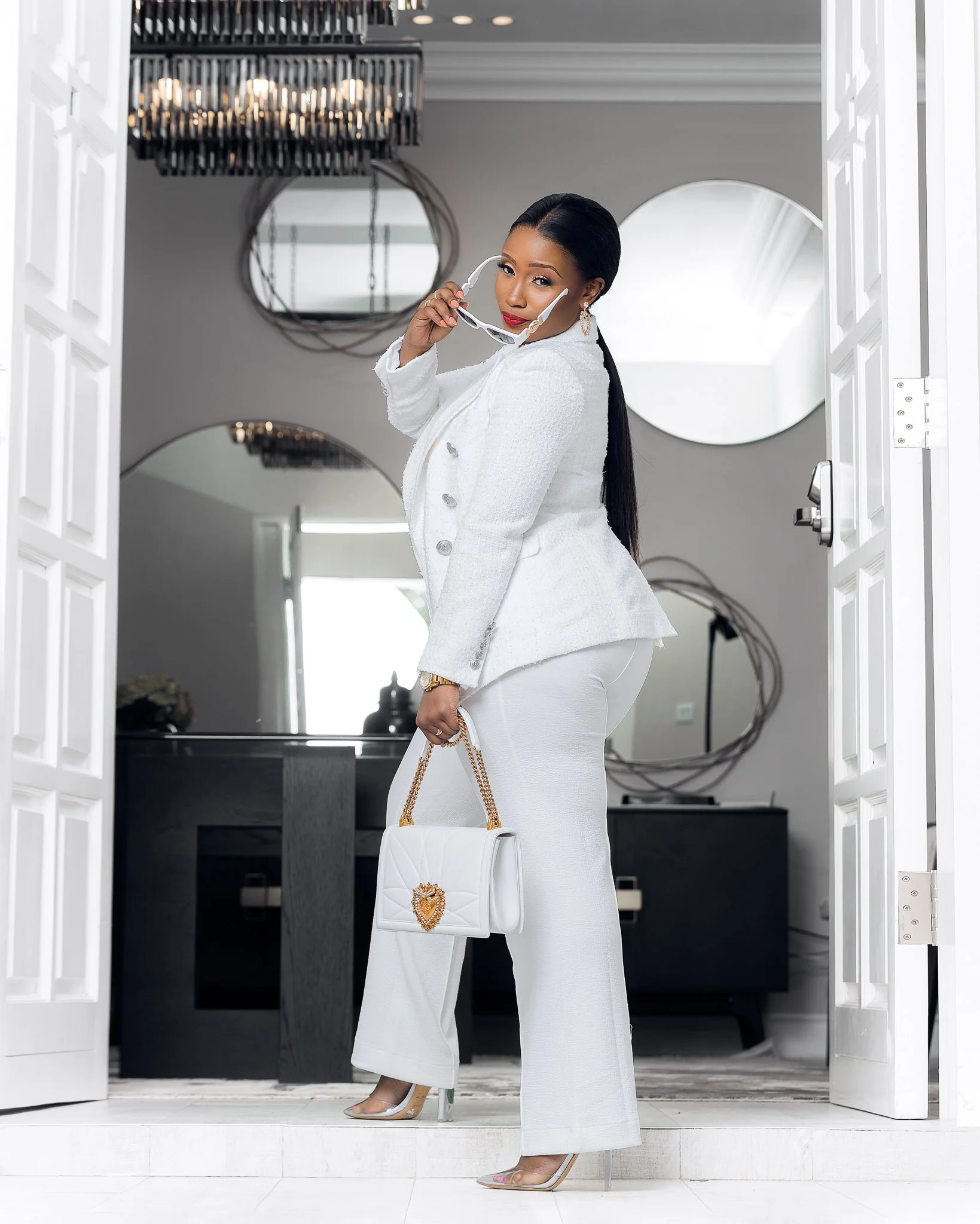 A woman dressed in a white suit holding sunglasses in one hand and a white handbag with a gold chain in the other, standing in a doorway in a modern, elegantly decorated room.