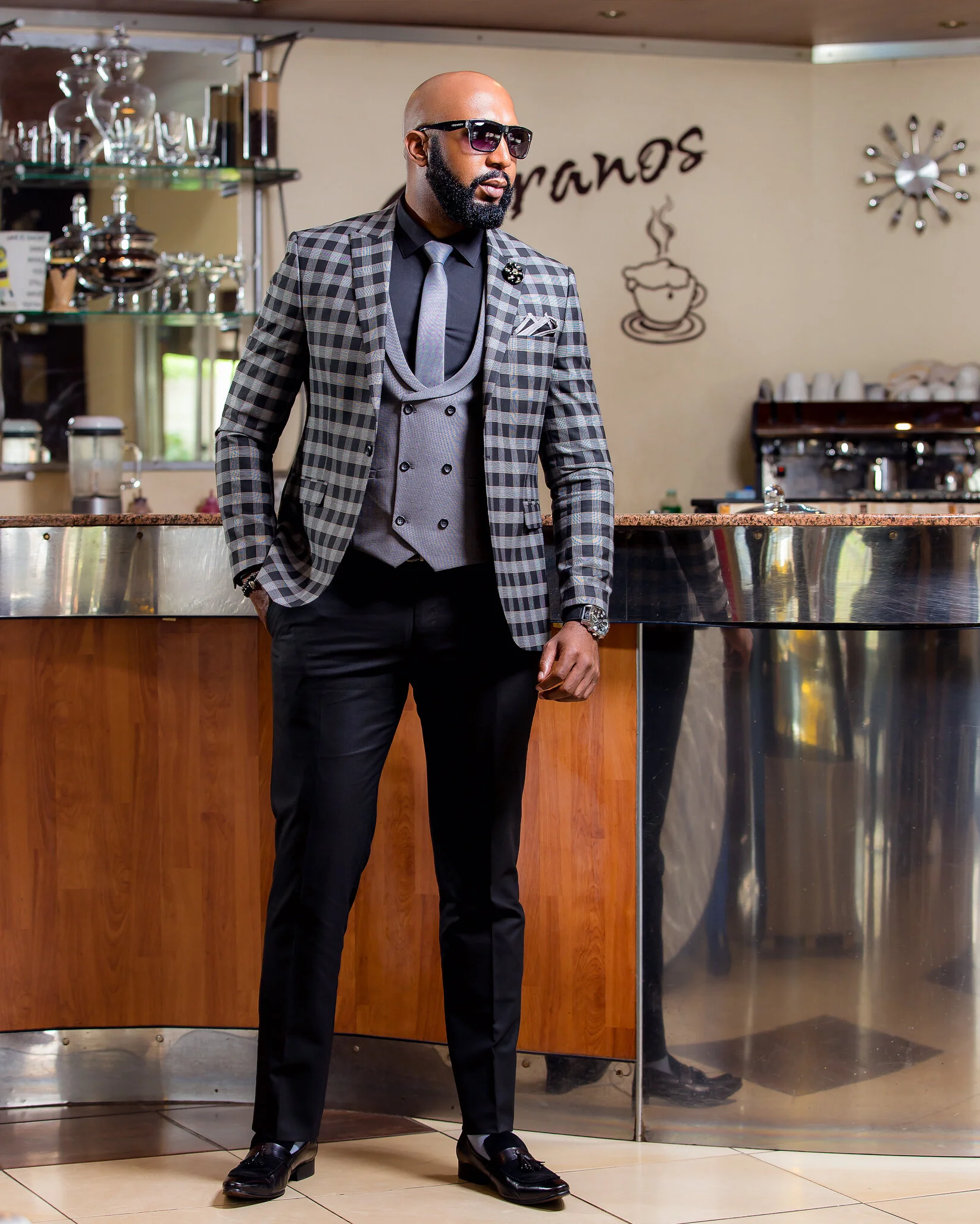 A man in a stylish checkered blazer, gray vest, black trousers, black shoes, sunglasses, and a watch stands in a cafe or restaurant near the counter.