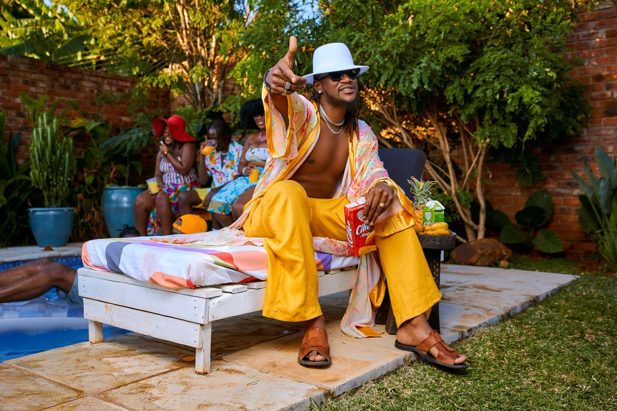 Man sitting on poolside lounge chair wearing yellow pants, a colorful open shirt, sunglasses, a white hat, and sandals, holding a box of Calypso drink, with a group of women relaxing in the background by the garden, some holding drinks and enjoying t