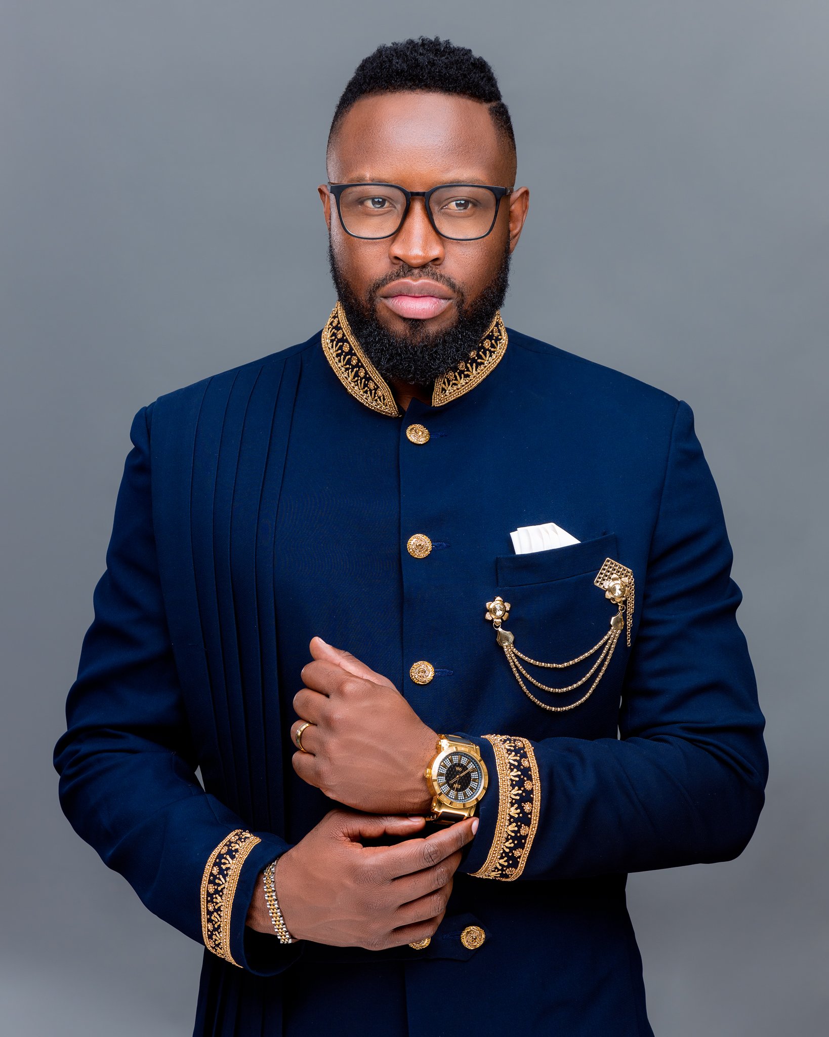 A man wearing a dark blue suit with gold embellishments, gold buttons, a gold chain, and a gold watch posing against a gray background.