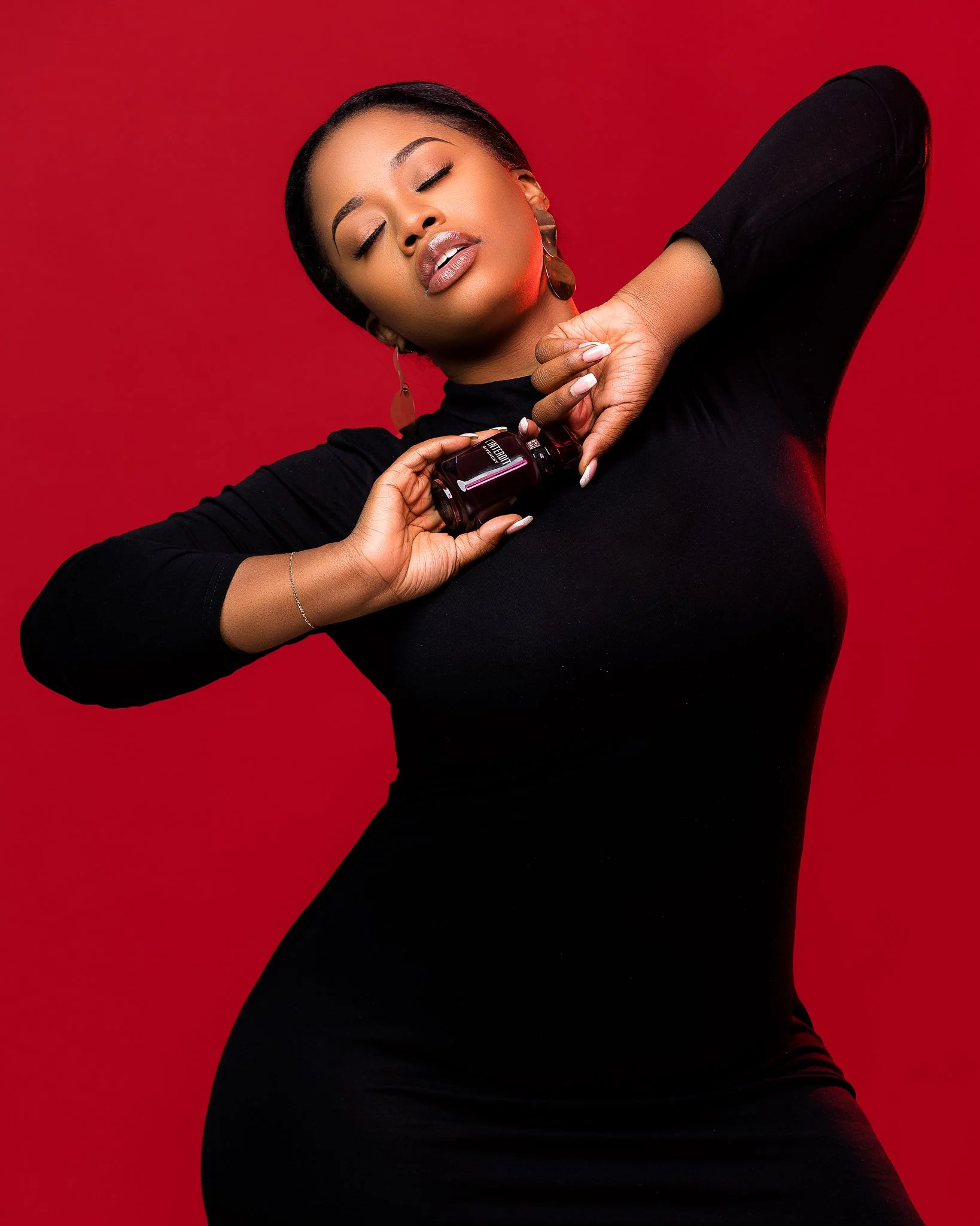A woman in a black dress holding a small bottle of nail polish on a red background.