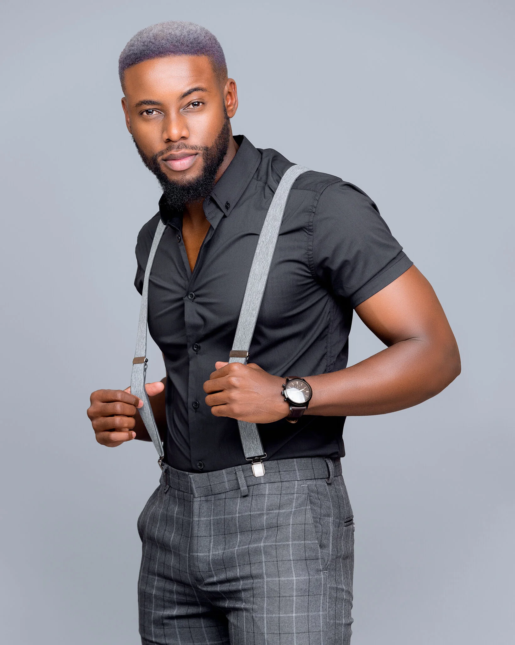 A young man with a neatly groomed beard and styled hair, wearing a black short-sleeve button-up shirt, gray plaid trousers, gray suspenders, and a black watch, posing against a plain gray background.
