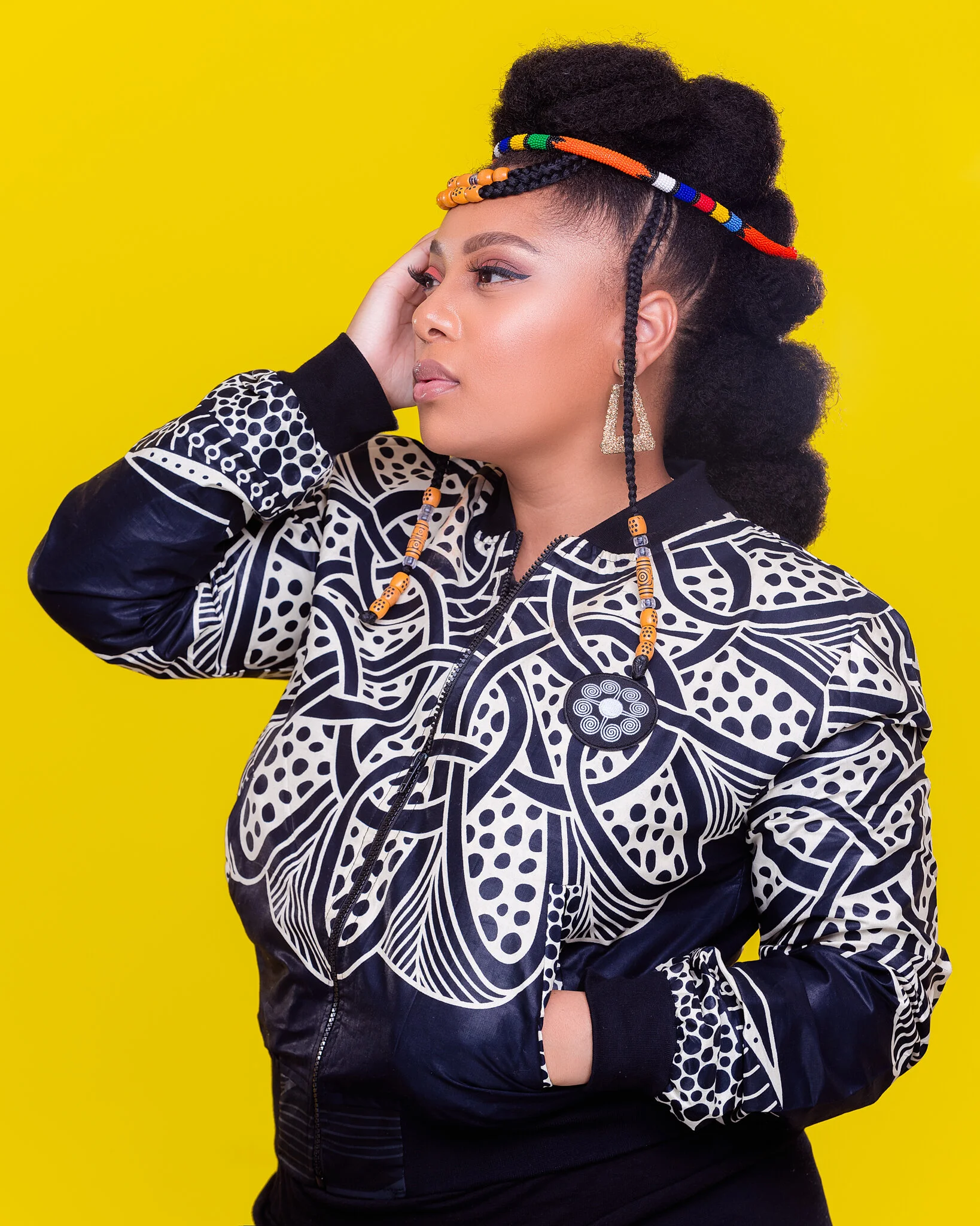 Portrait of a woman with styled hair, wearing a patterned jacket, colorful beaded headband, geometric earrings, and a large decorative necklace against a yellow background.