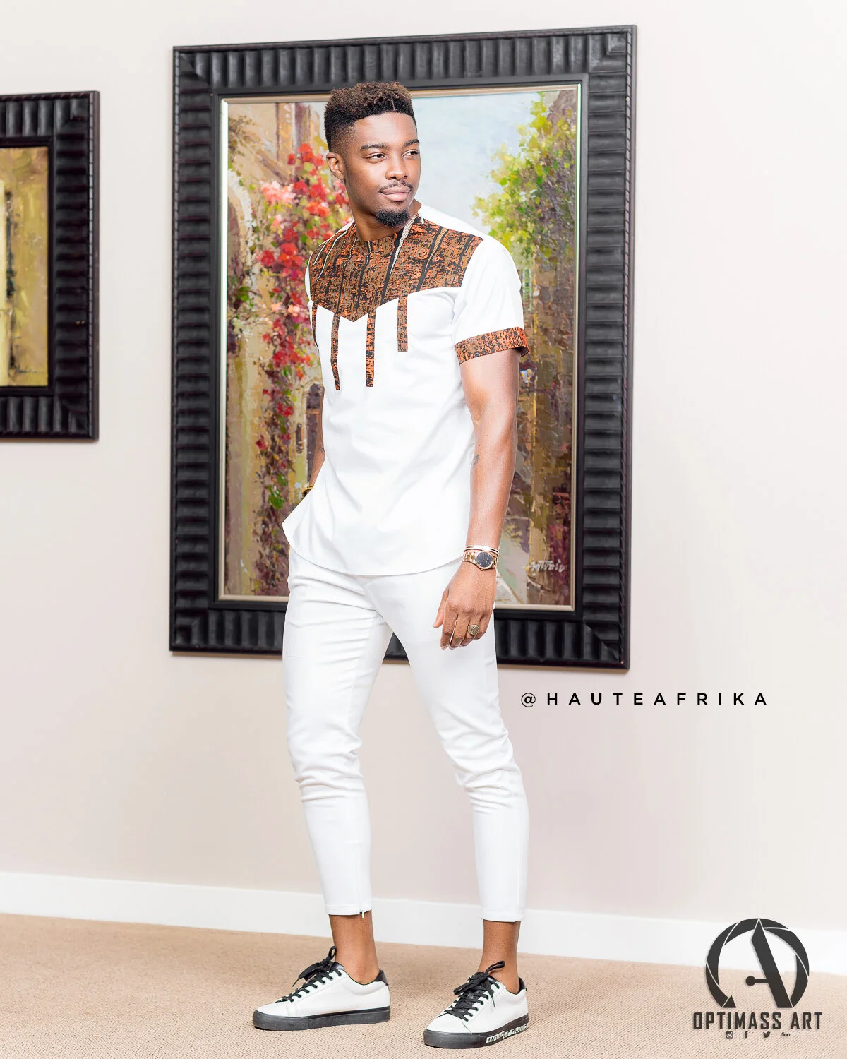 A man in a white outfit with African-inspired design accents stands in an art gallery, looking to the side, with framed artwork behind him.