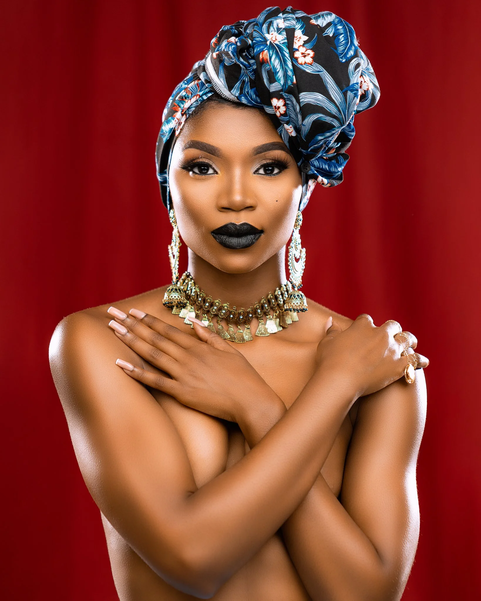 A woman with dark lipstick and eye makeup wears a patterned headwrap, multiple necklaces, and earrings, standing against a red background.