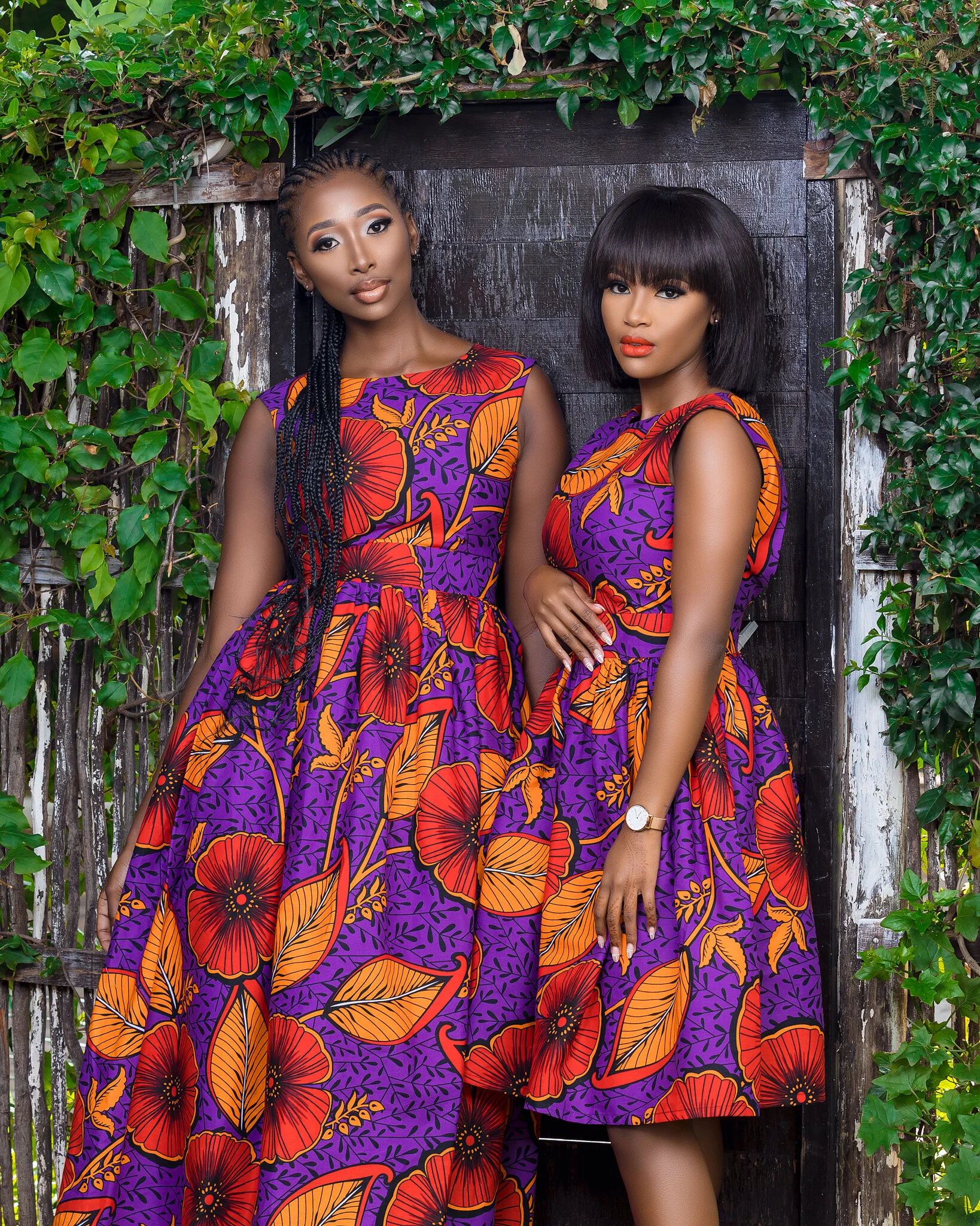 Two women in colorful purple, orange, and red floral dresses standing against a black wooden background with green leafy vines surrounding them.