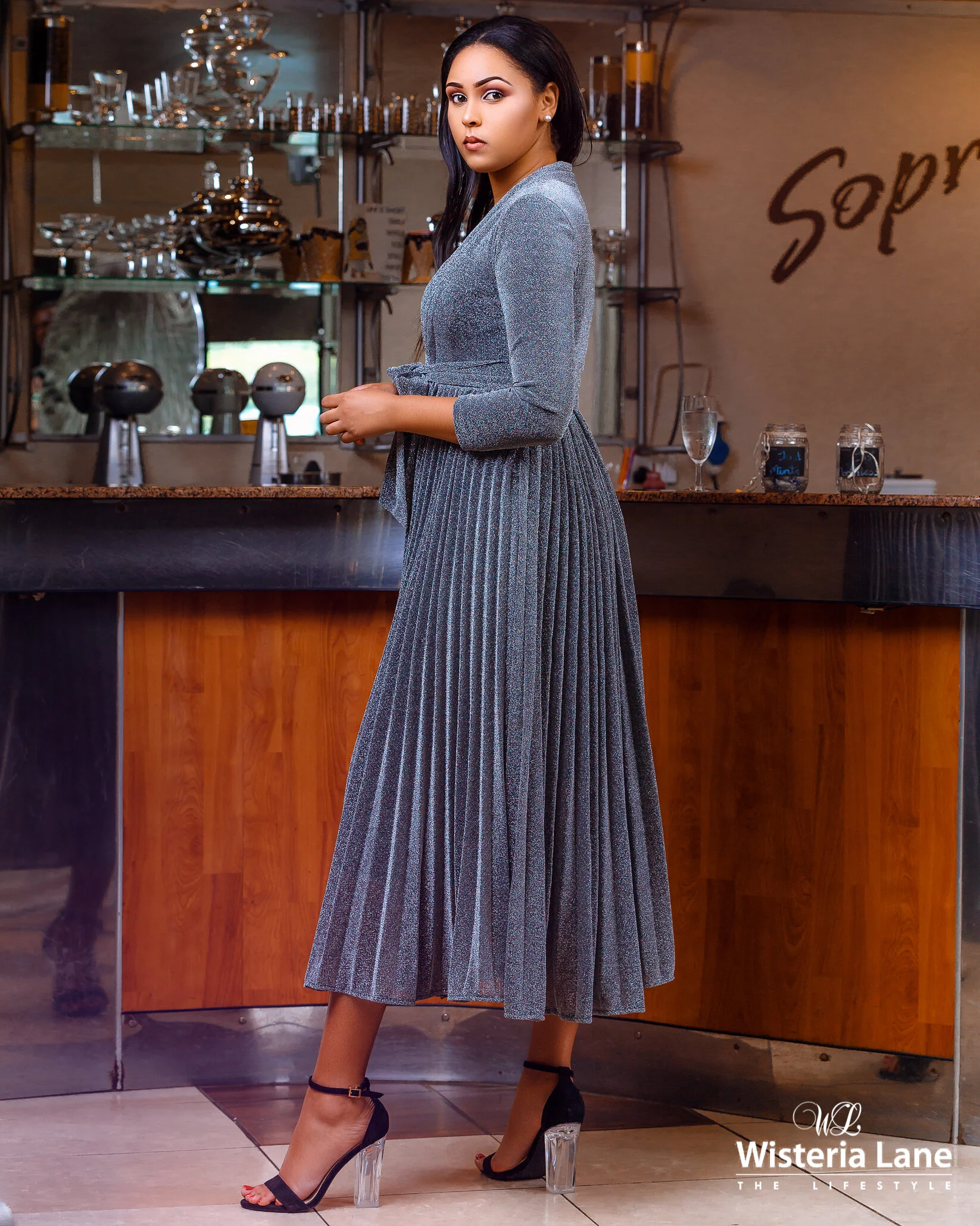 A woman wearing a sparkly gray dress with a pleated skirt and black high heels, standing in a stylish cafe or restaurant with a bar counter in the background.