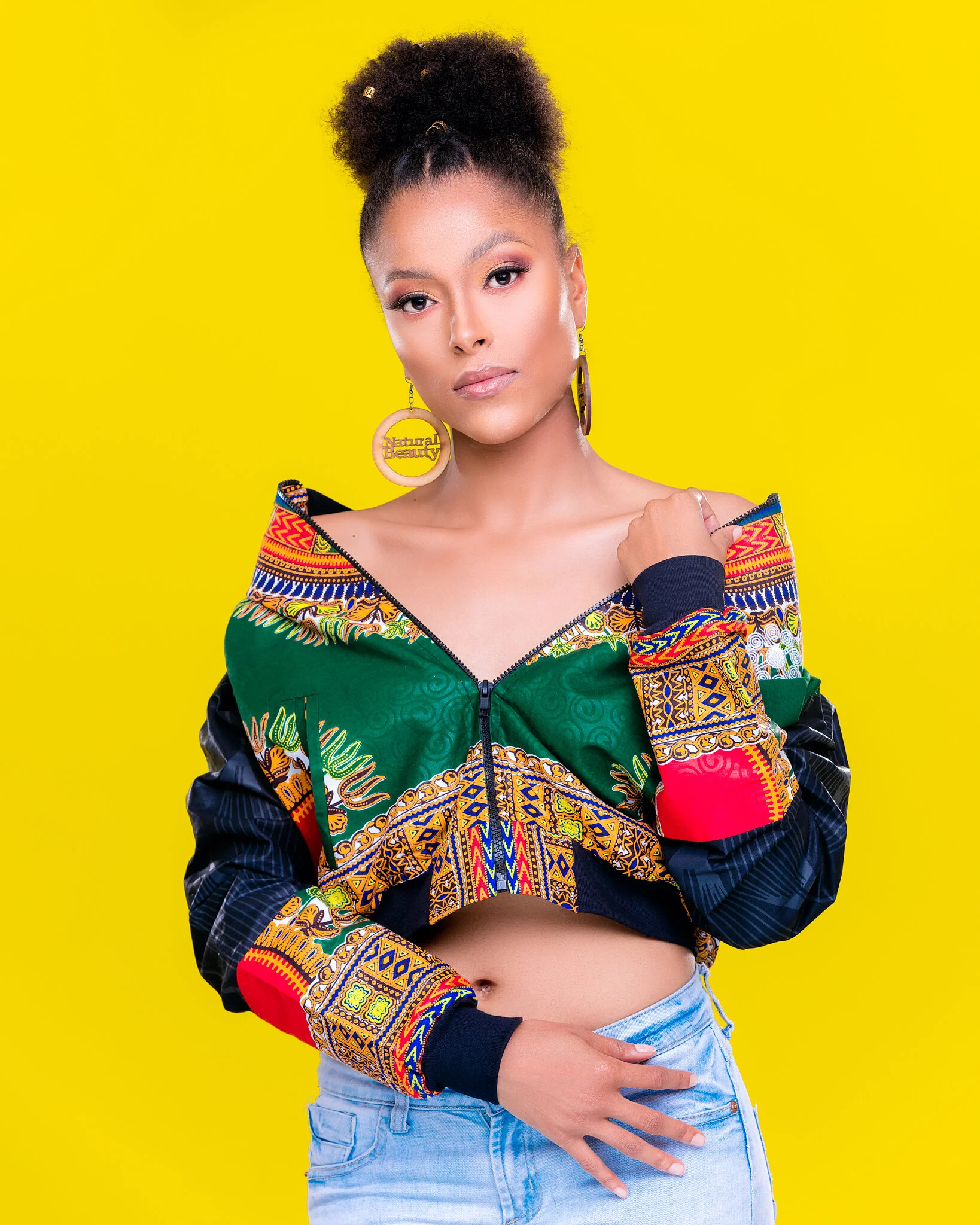Young woman with styled curly hair in a high puff, wearing a colorful African print jacket over a black top, and light blue jeans, standing against a bright yellow background.