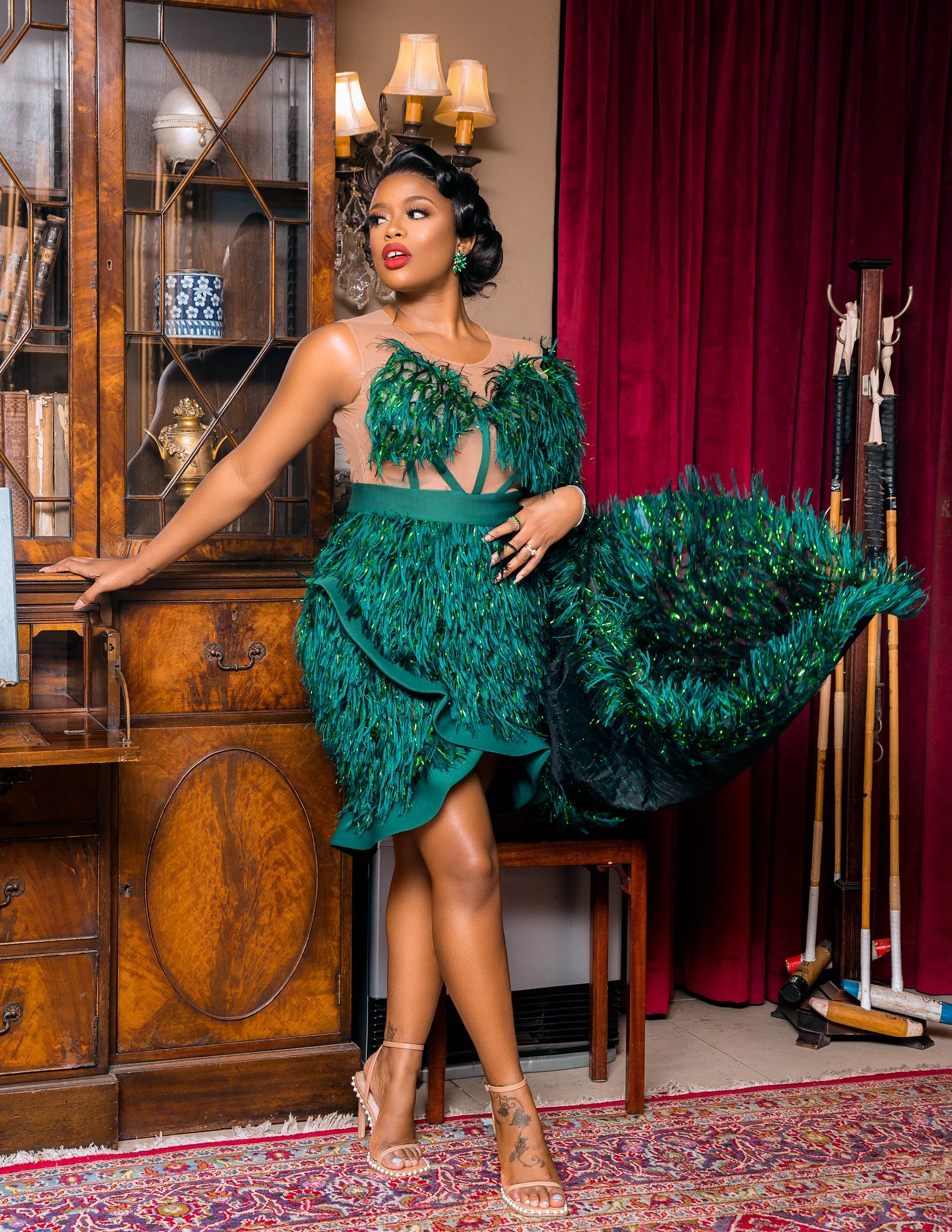 A woman in a green feathered dress standing next to a wooden cabinet, with red curtains and golf clubs in the background.