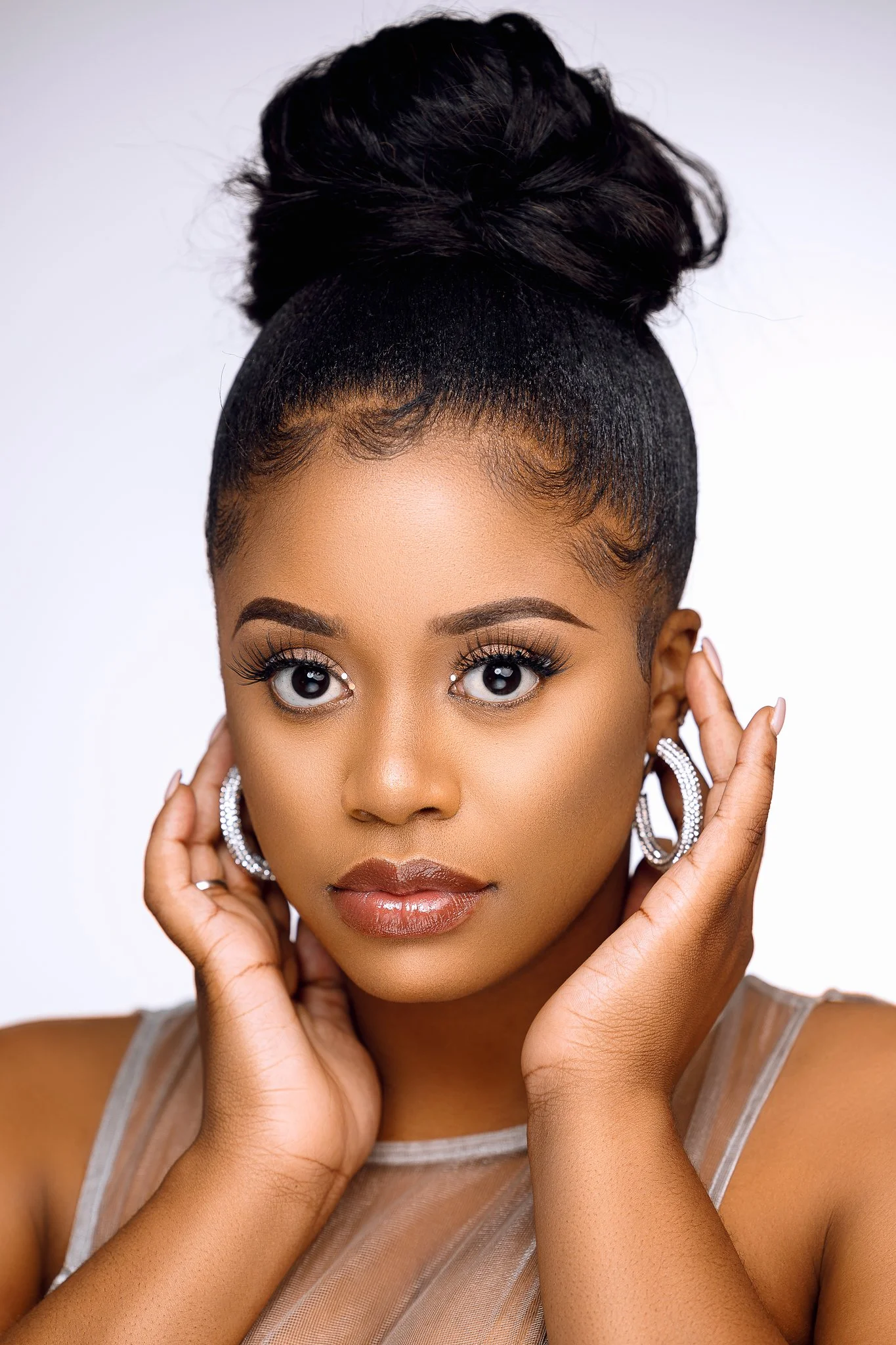 Close-up portrait of a young woman with styled hair in an updo, wearing large hoop earrings and a sheer top, touching her face gently.