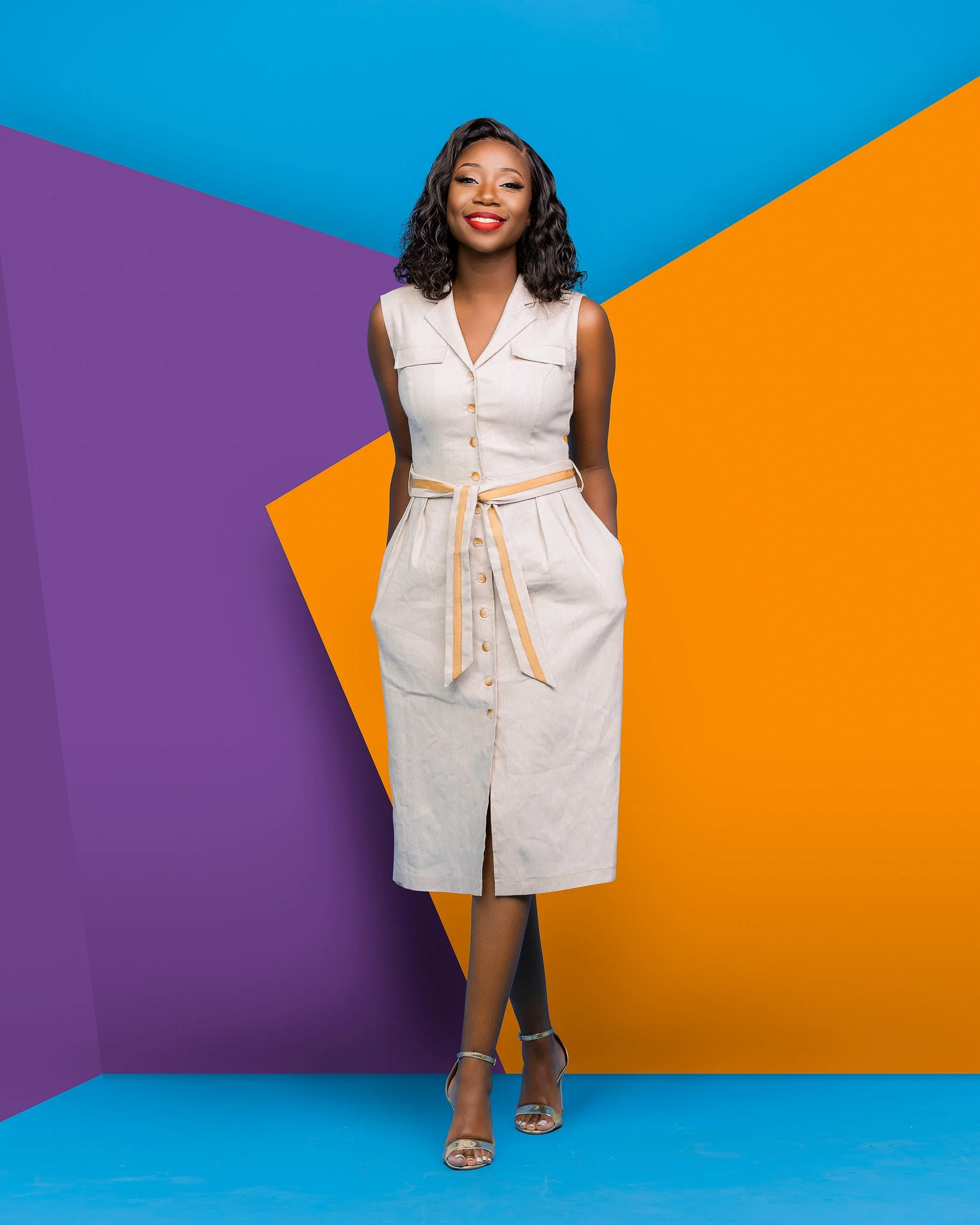A woman with dark curly hair and red lipstick smiling, wearing a sleeveless beige dress with buttons and a tied waist, standing against a colorful geometric background with blue, purple, and orange sections.