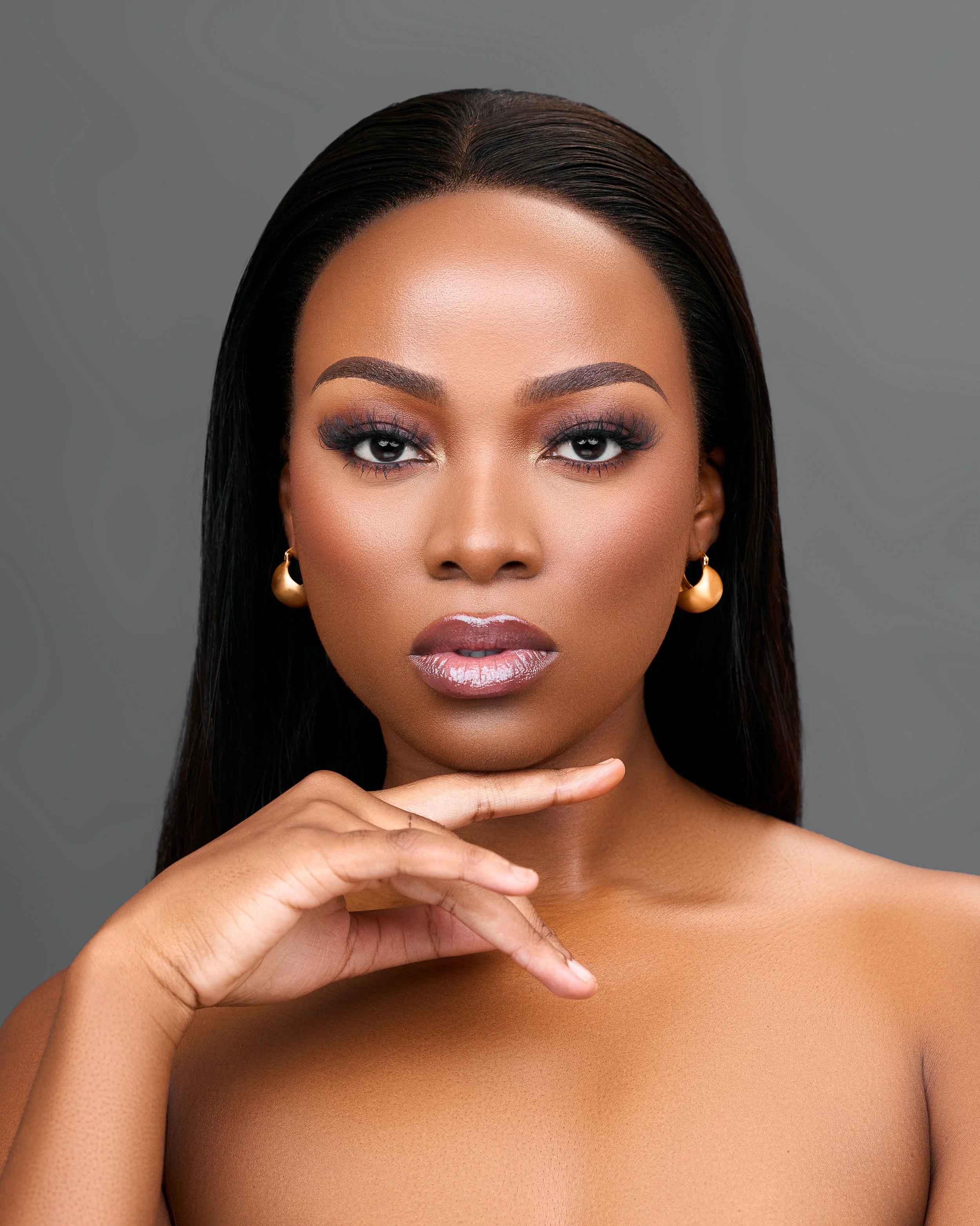 Close-up of a woman with dark hair, gold earrings, and makeup. She is looking directly at the camera with a serious expression, her left hand gently touching her chin.