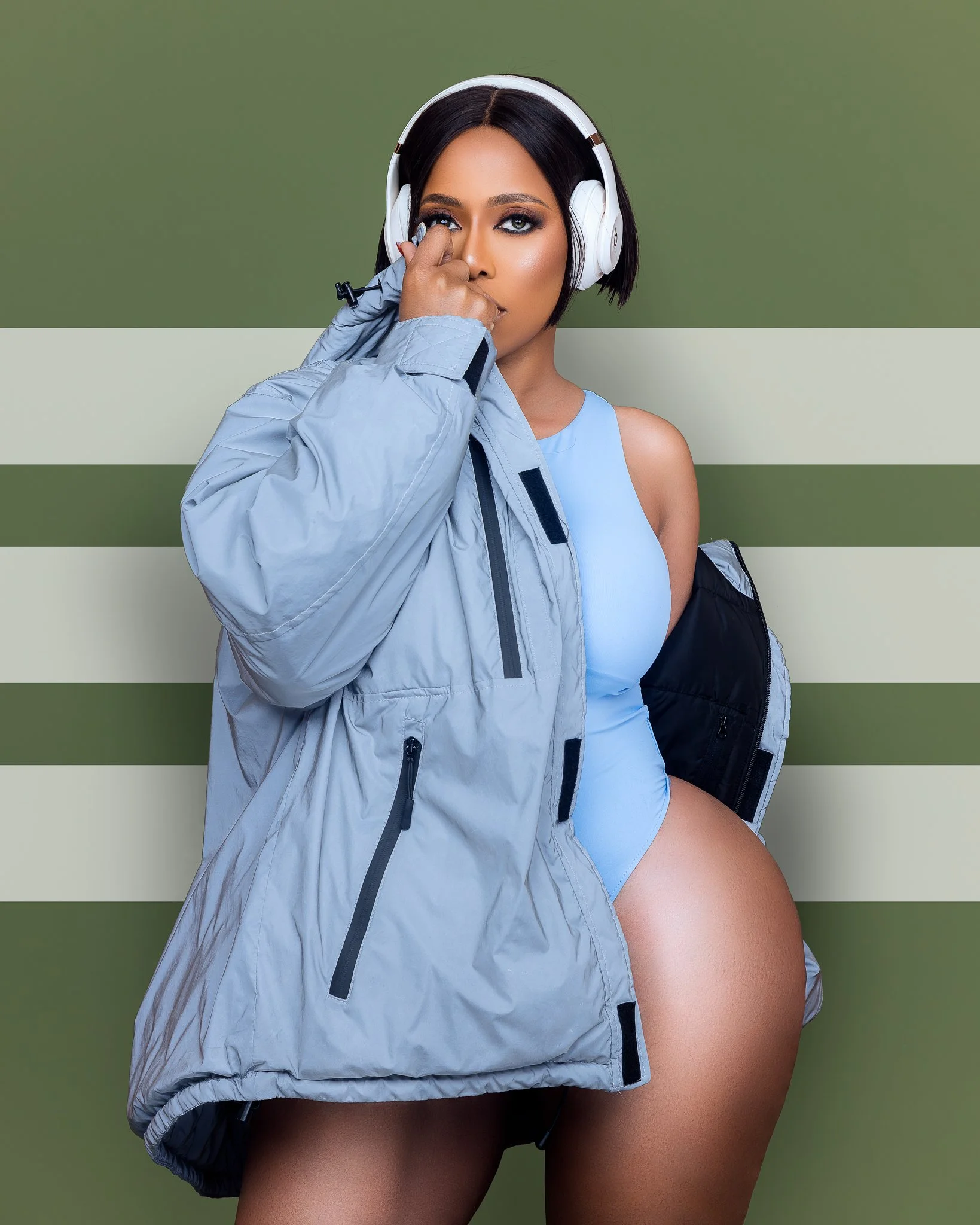 A woman with short black hair wearing a light blue sports outfit, headphones, and a gray jacket, standing in front of a green-striped background, with a black backpack over her shoulder, holding her jacket with one hand, and touching her face with he