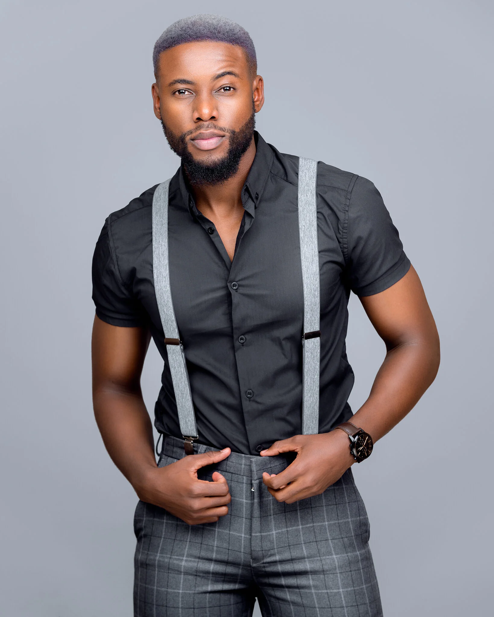 A stylish man with a beard and short gray hair wearing a black button-up shirt with gray suspenders, gray plaid pants, and a wristwatch against a plain gray background.