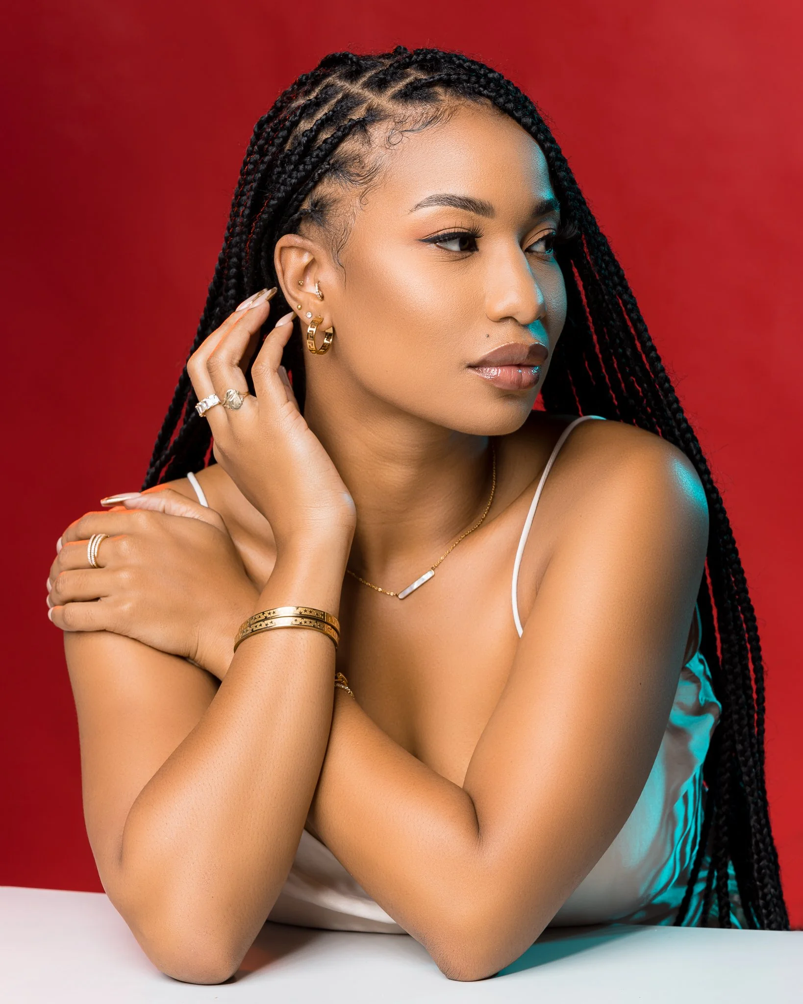 A woman with long braided hair wearing jewelry posing against a red background.