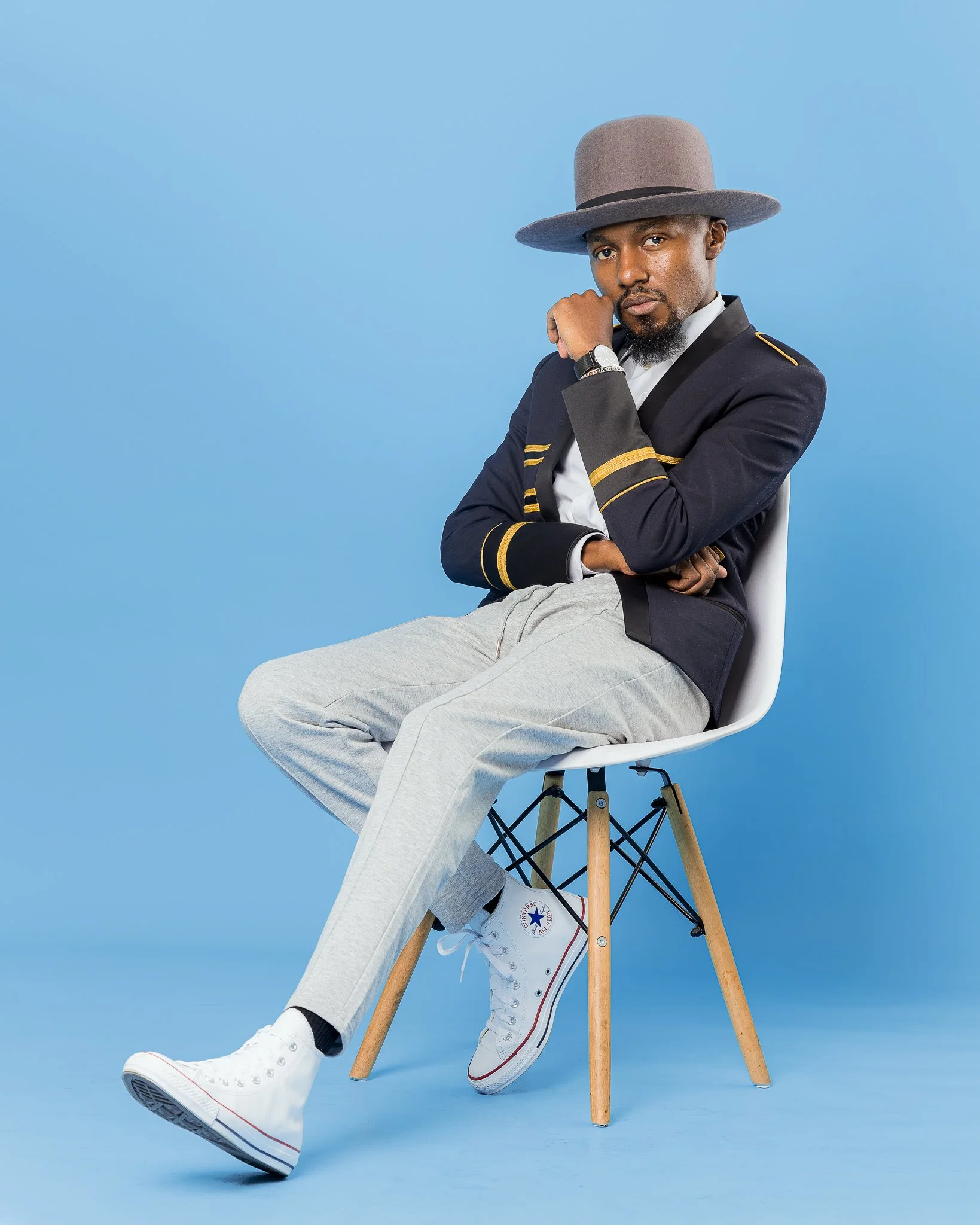 A man sitting on a white chair with wooden legs against a blue background, wearing a wide-brimmed gray hat, a navy blazer with gold trim, light gray pants, white high-top Converse sneakers, and a wristwatch, with his left arm crossed and his right ha