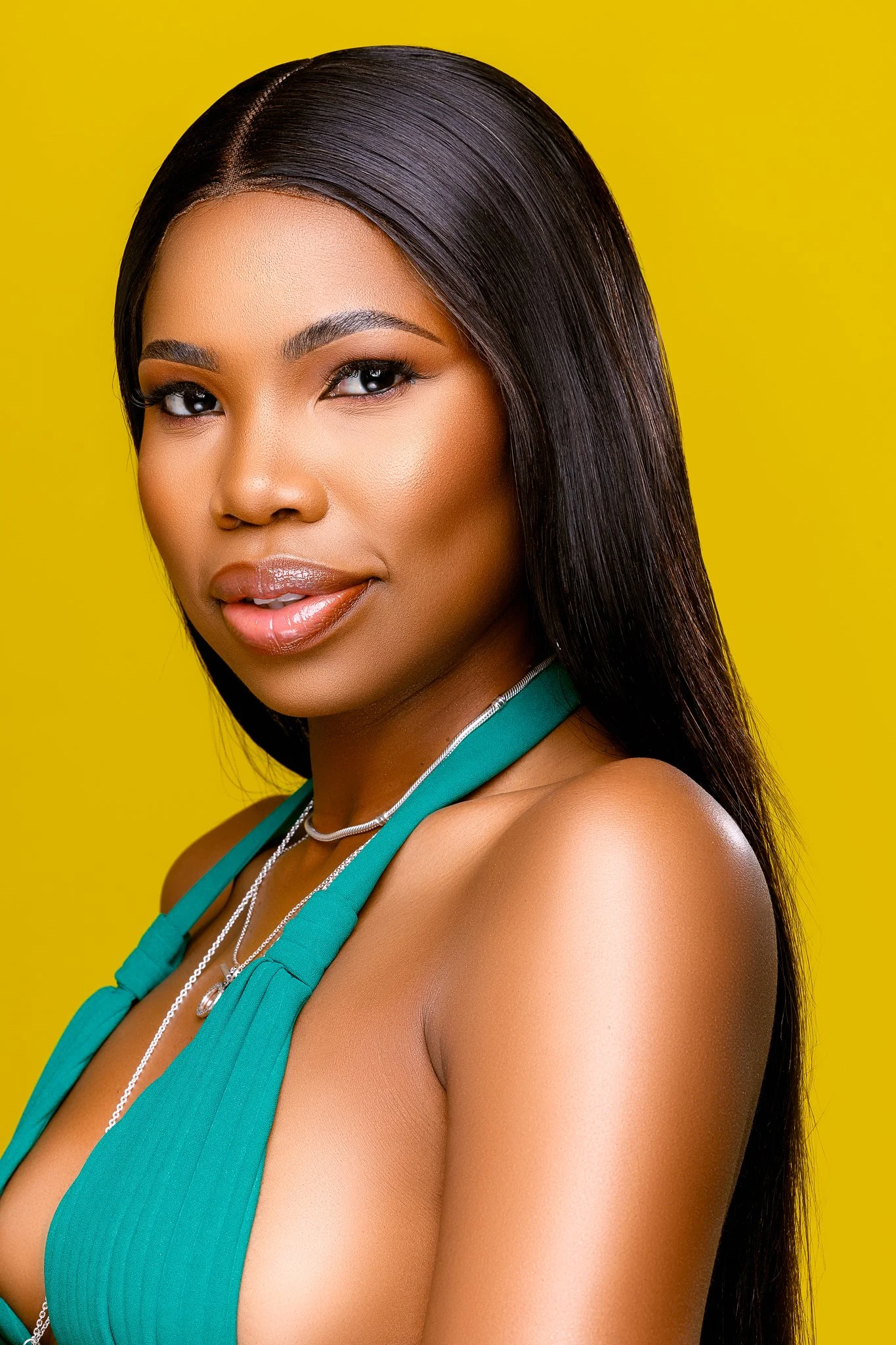 Close-up of a woman with long dark hair, smiling, wearing makeup, a teal top, and layered necklaces, against a yellow background.