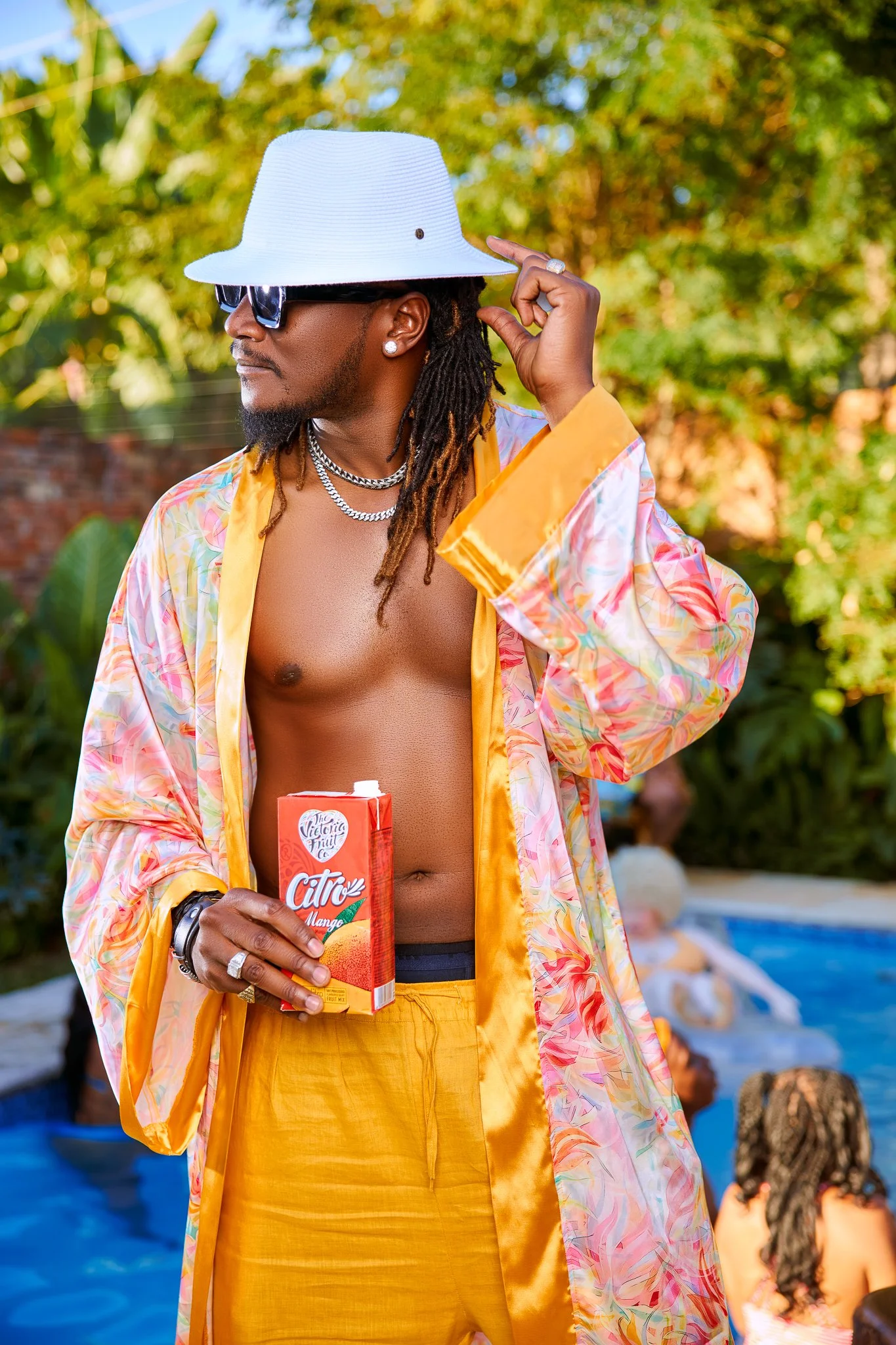 A man with dreadlocks wearing sunglasses, a white wide-brimmed hat, and yellow pants, standing by a pool holding a juice box, with other people relaxing in the background.