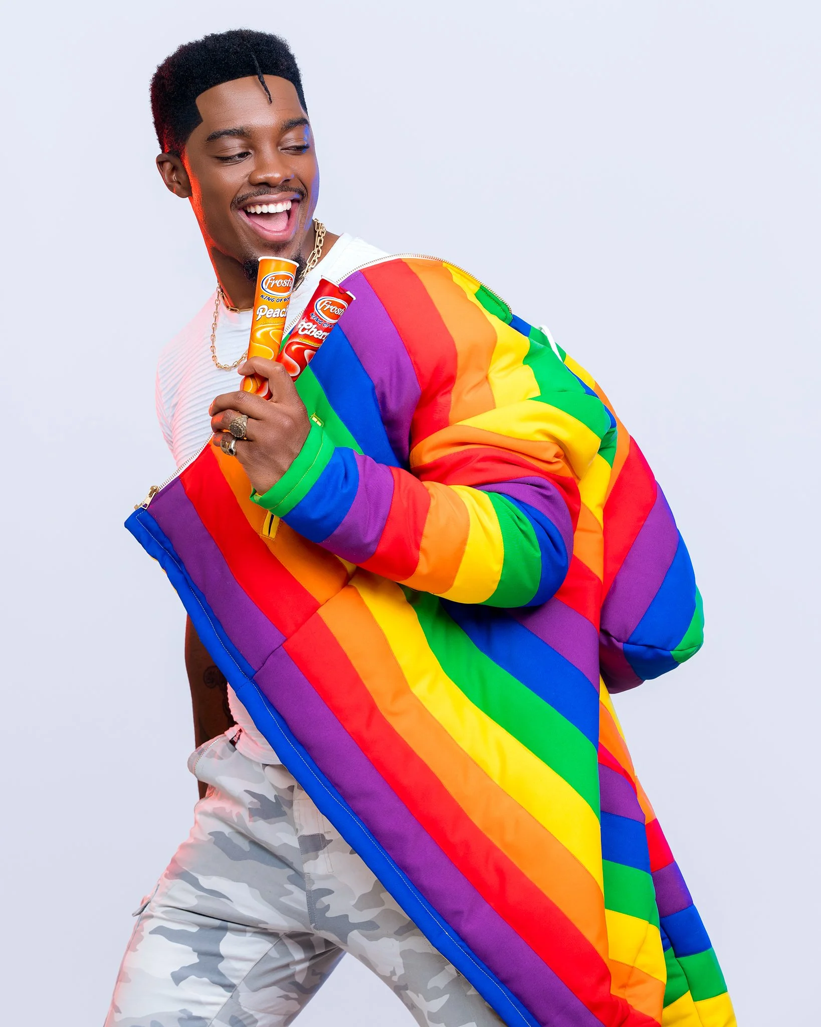 A smiling man in a rainbow-colored jacket holding fruity flavored ice pops, standing against a light background.