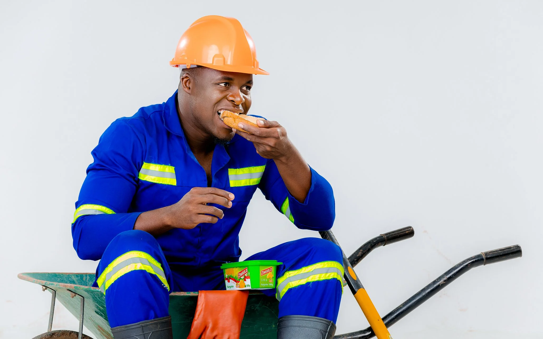 A man in construction worker attire, wearing a blue uniform with reflective yellow stripes and an orange safety helmet, sitting on a wheelbarrow, eating a snack, with two small containers of margarine in front of him.