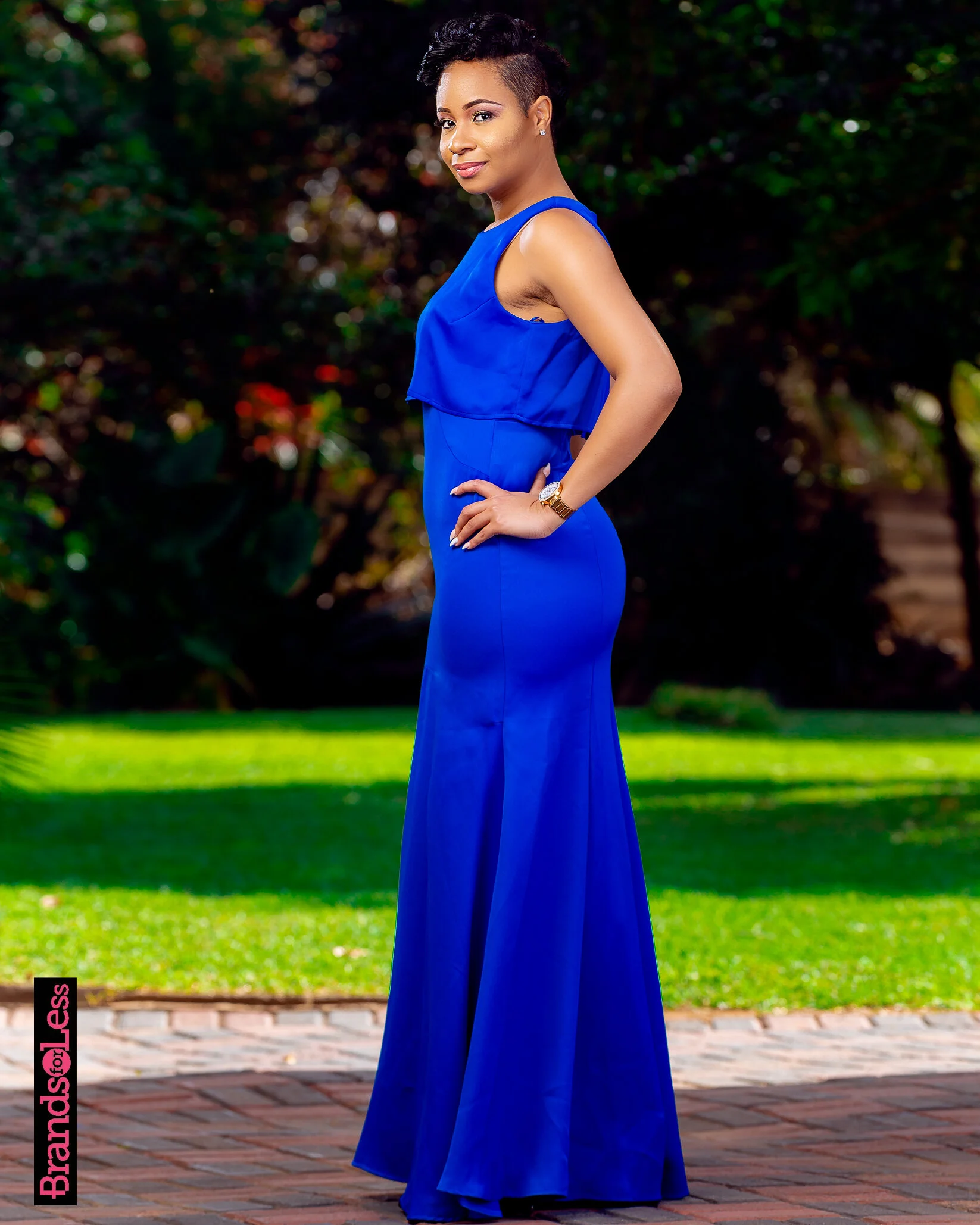 A woman in a bright blue sleeveless dress stands outdoors on a brick path with greenery in the background, looking at the camera with a slight smile.