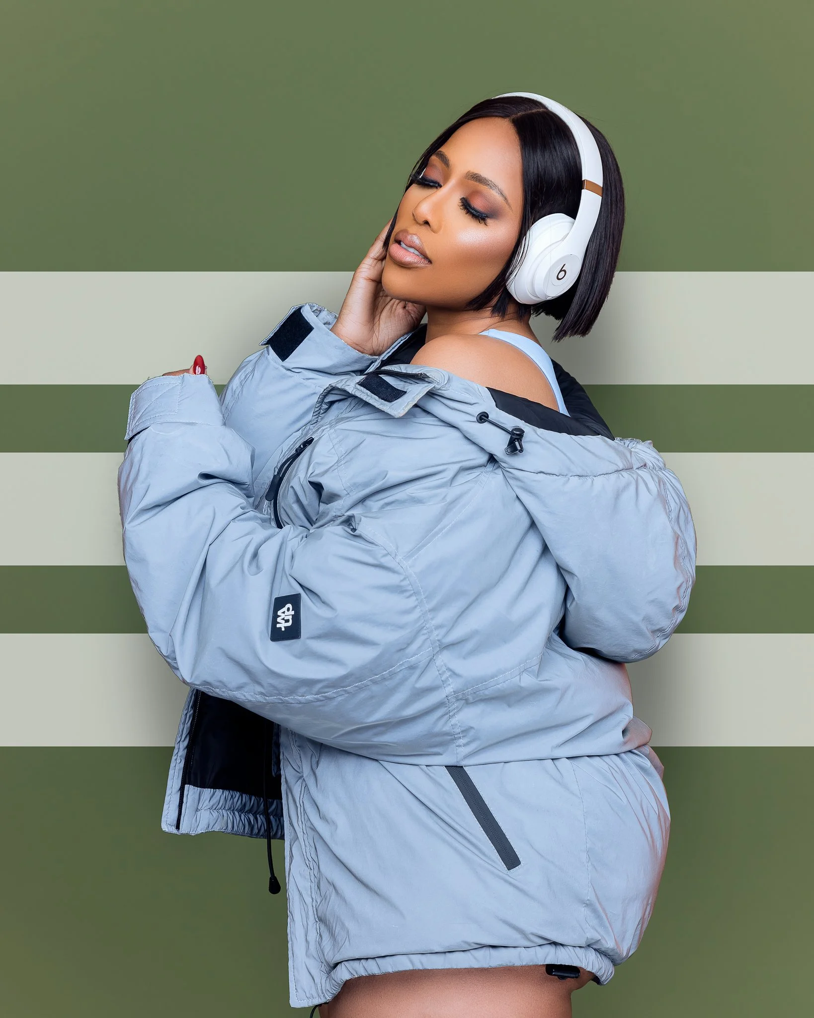 A woman with short black hair wearing large white headphones, a blue tank top, and a light gray puffer jacket, standing against a green and beige striped background, with her eyes closed and a relaxed expression.