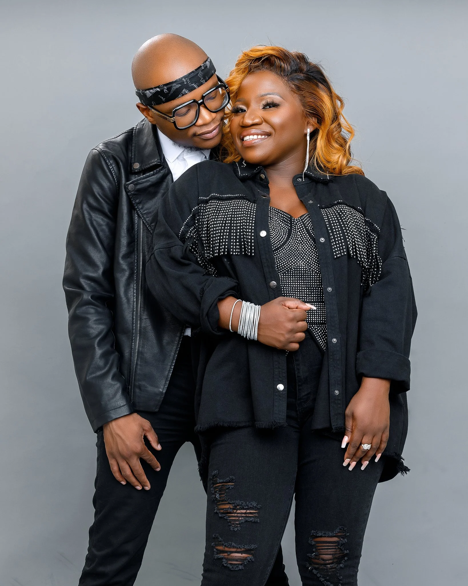 A stylish couple dressed in black clothing, standing close together against a gray background, smiling and posing for the camera.