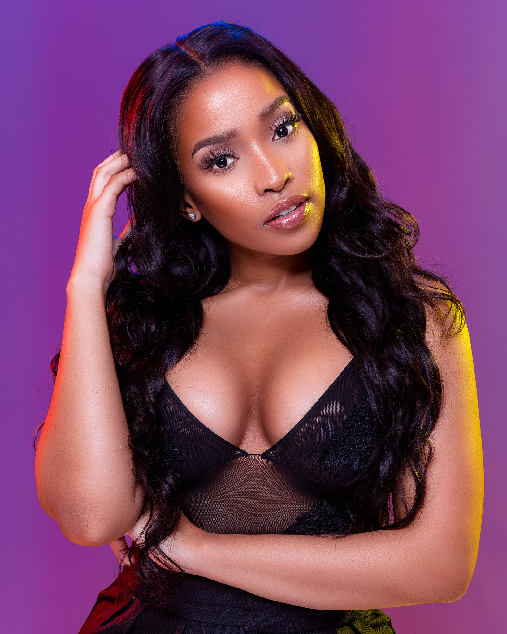 Portrait of a woman with long curly black hair, wearing a black lace top, posing with her hand near her head against a purple background.