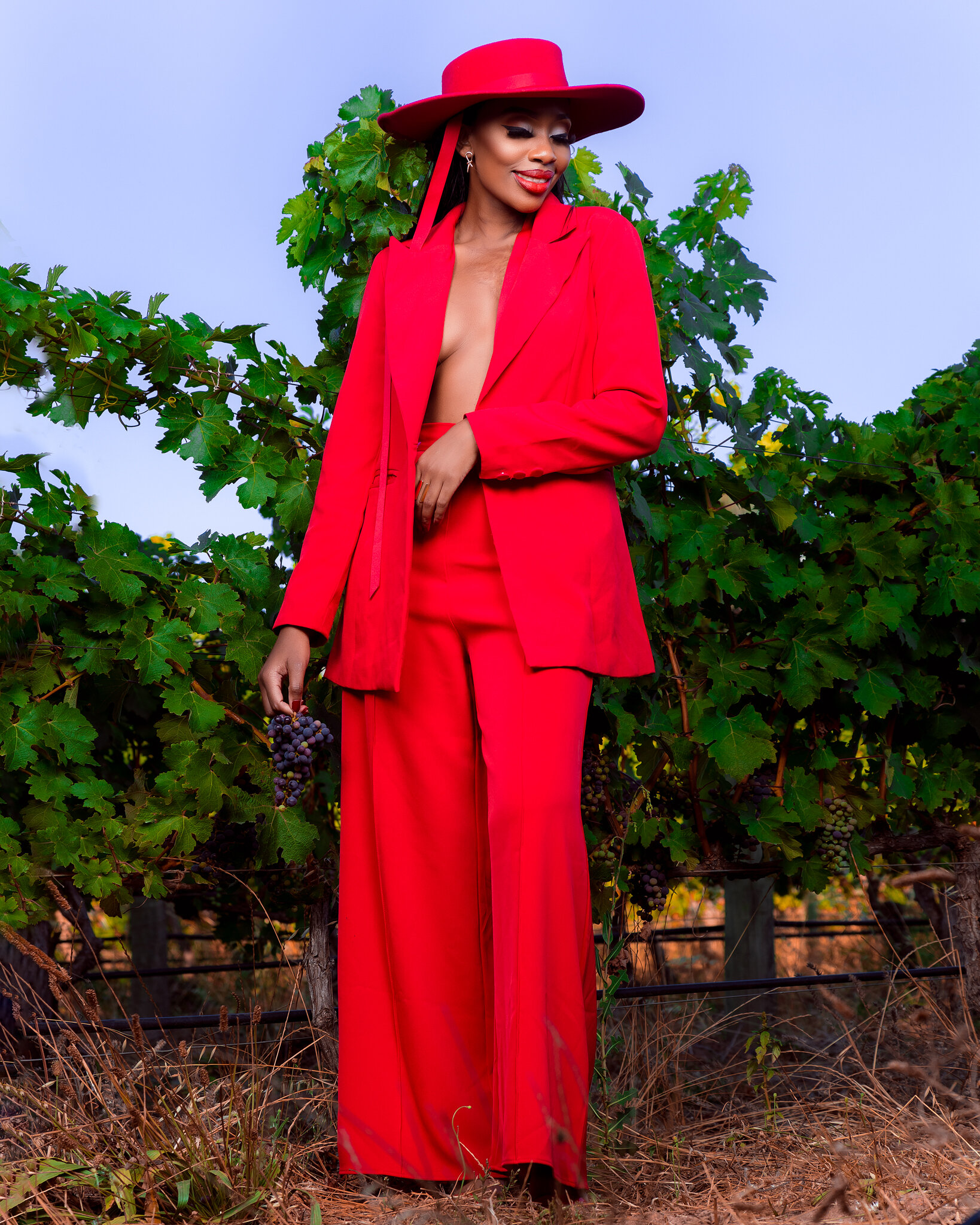 A woman dressed in a bright red suit and wide-brimmed hat stands in a vineyard, holding a bunch of grapes.